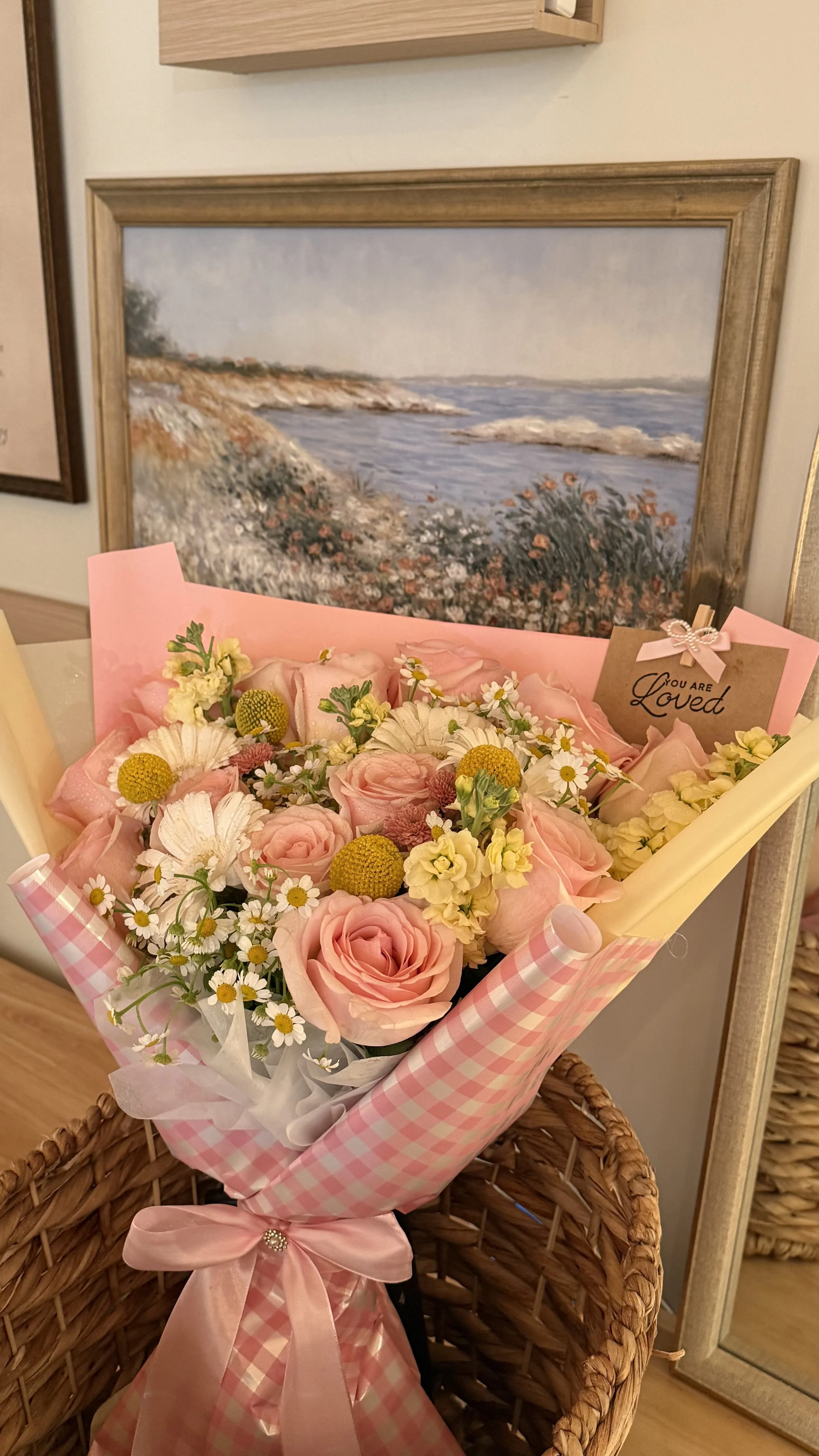 A pink checkered gift bouquet with roses, daisies, and other flowers on a wicker chair, in front of a landscape painting of the seaside with rocky cliffs and water.
