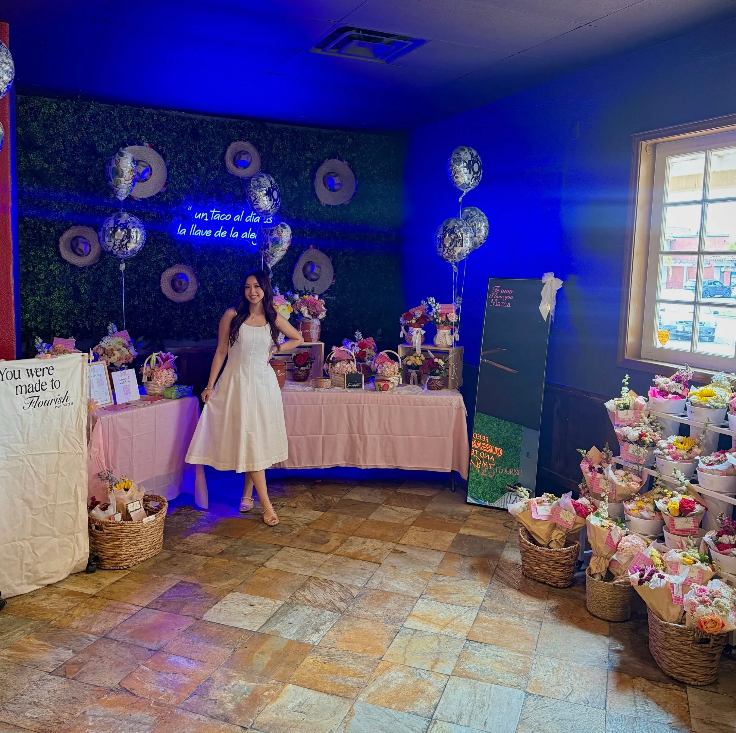 Indoor floral display at a celebration or event, with a woman in white dress standing in front, decorated with bouquets, balloons, and signs, near a window letting in natural light.