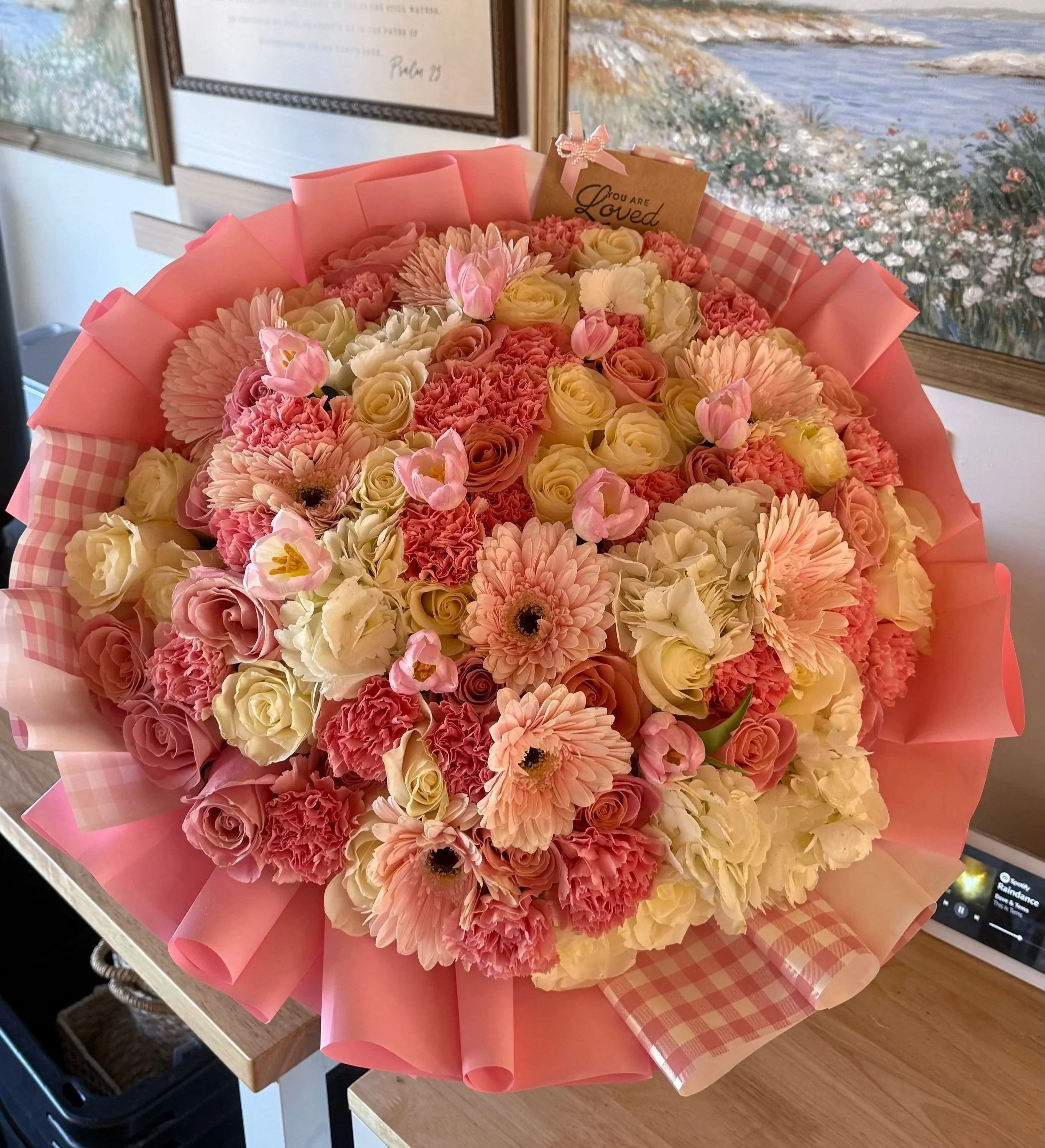 A vibrant bouquet of pink, cream, and pastel-colored flowers wrapped in pink paper sits on a wooden table with a tablecloth, with framed artwork and a small sign reading 'You are Loved' in the background.