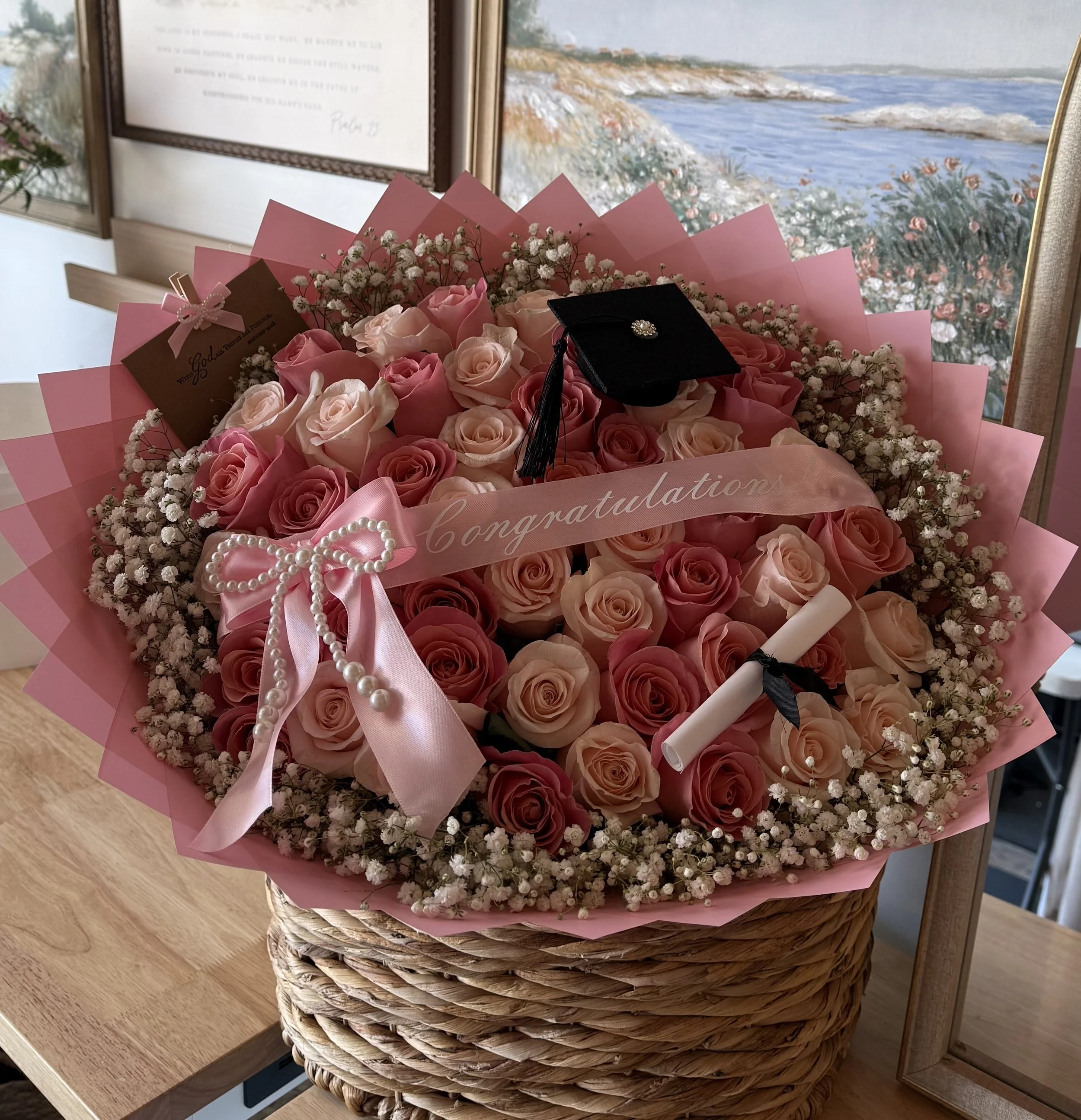 A bouquet of pink roses and small white flowers inside pink wrapping paper, placed in a woven basket. A graduation cap with a decorative brooch and a pearl necklace are on top of the bouquet. There are two greeting cards or notes among the flowers. T
