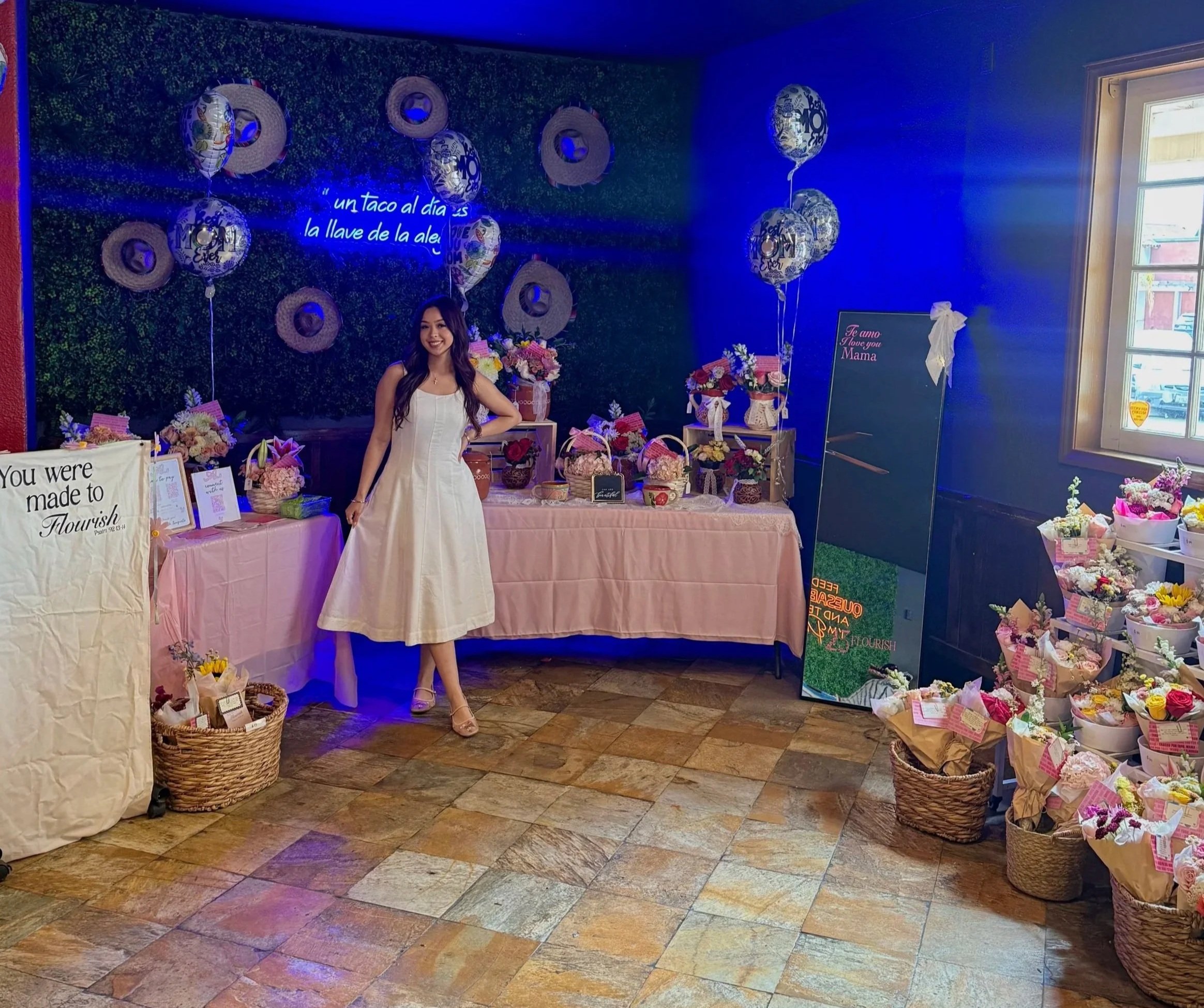 A young woman in a white dress standing next to a display of flowers and decorations inside a room with a green wall and sunlight coming through a window.