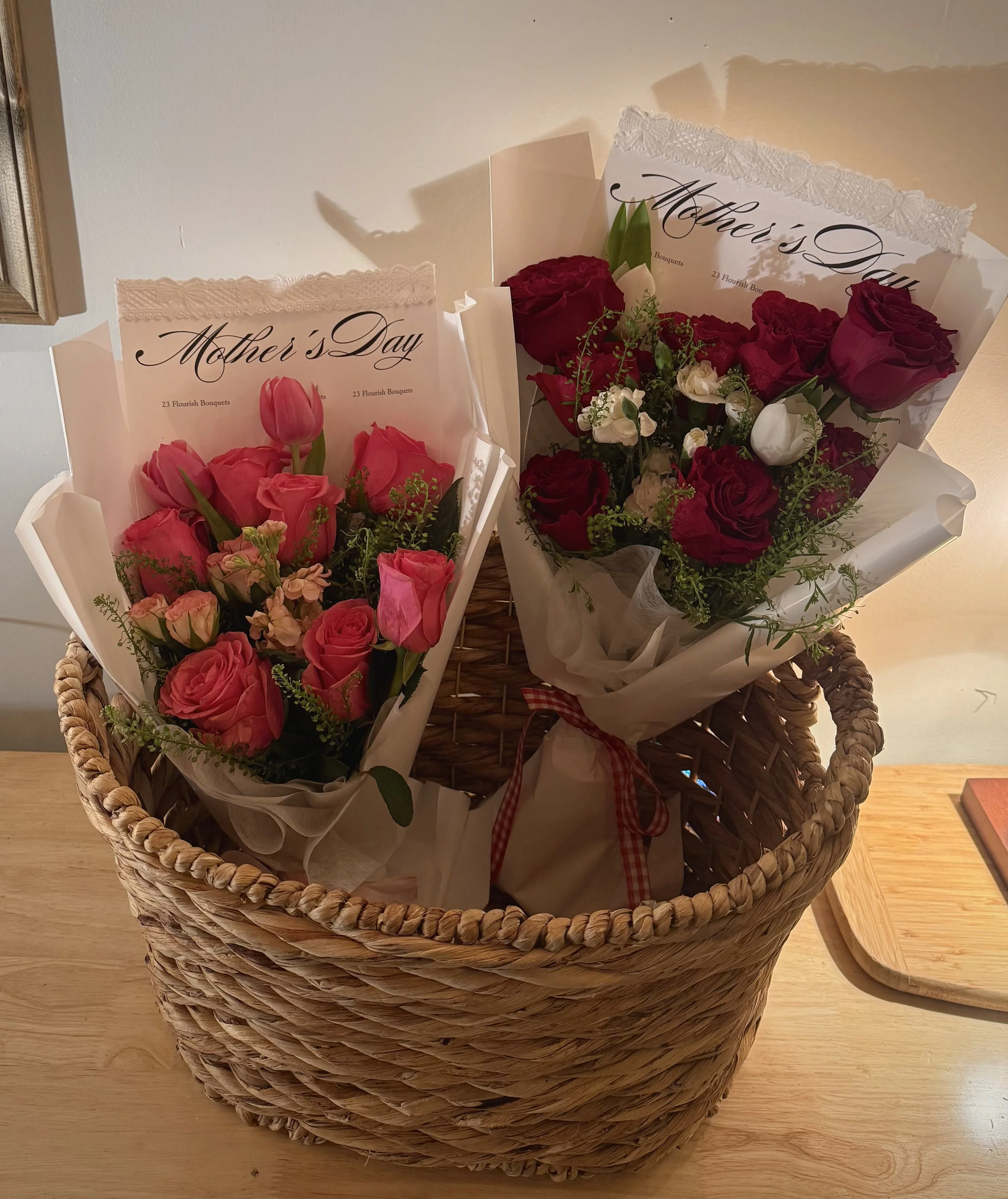 A wicker basket on a wooden surface contains bouquets of mixed pink, red, and white flowers with 'Mother's Day' cards.