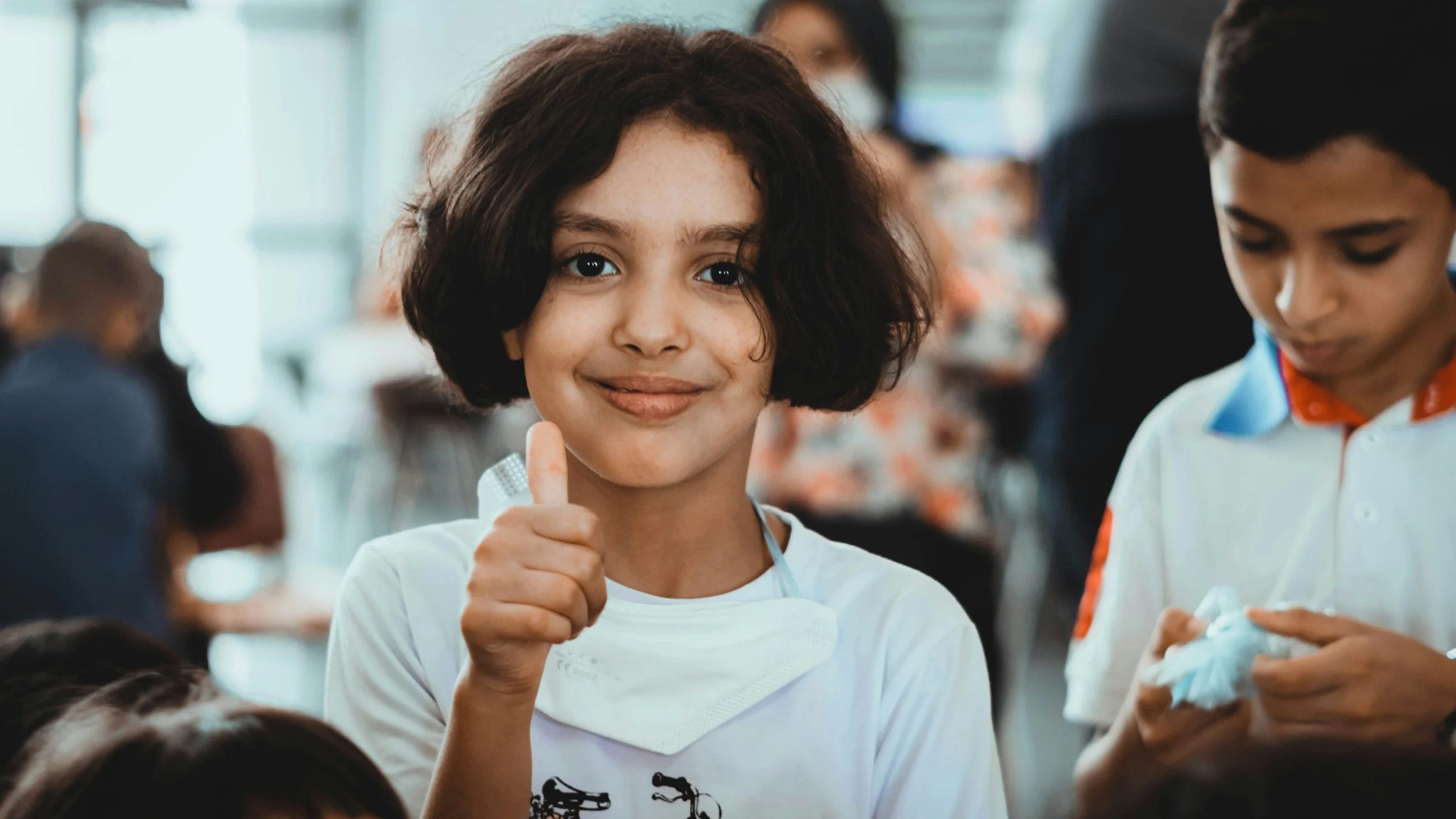 A young girl with short dark curly hair giving a thumbs up and smiling in a crowded indoor space.