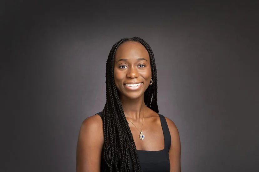 A smiling woman with long braided hair wearing a black sleeveless top and a necklace, against a plain dark background.