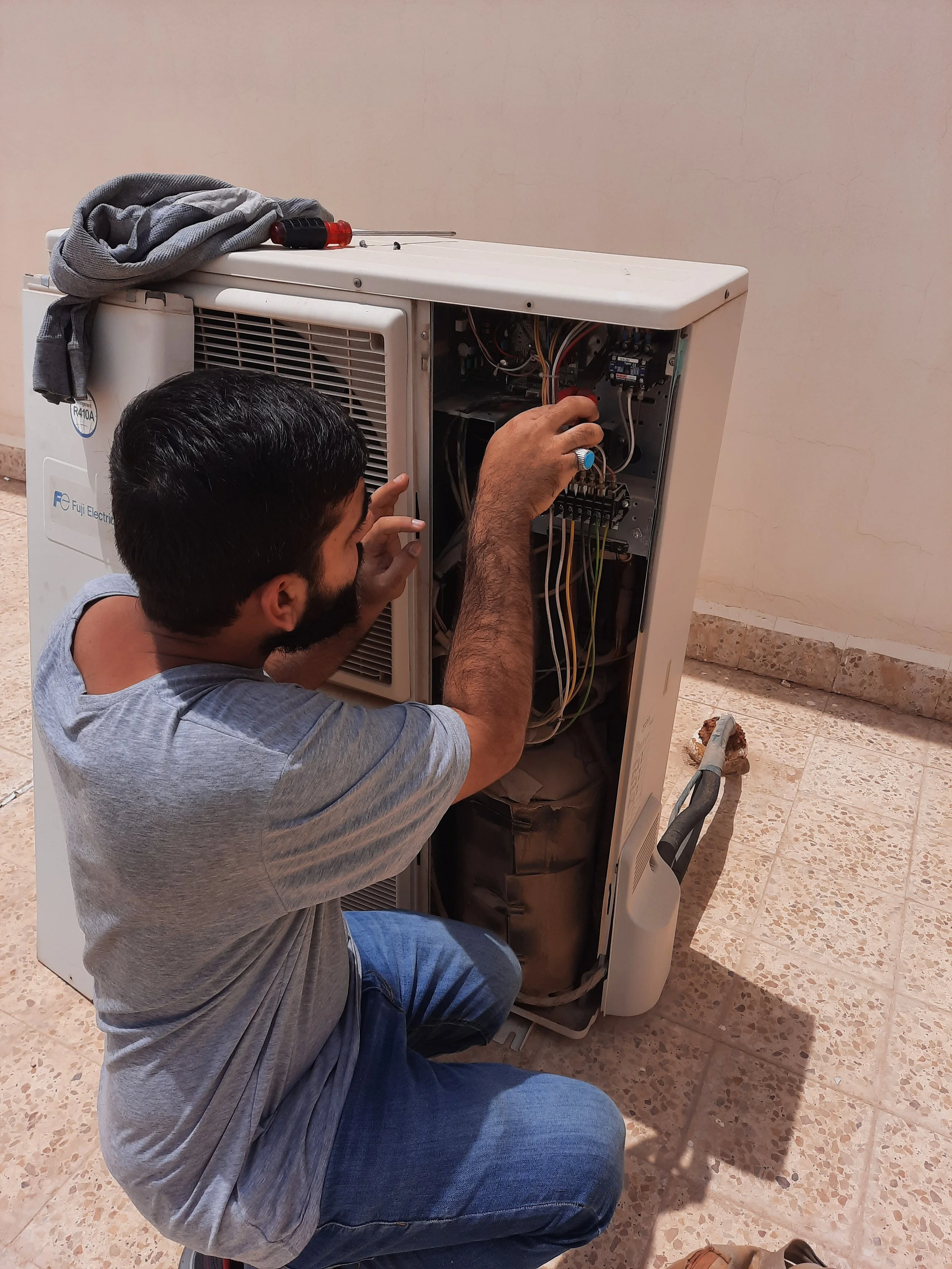 A technician repairing an air conditioning unit, working on the internal components with tools. The unit is outdoors on a tiled surface.