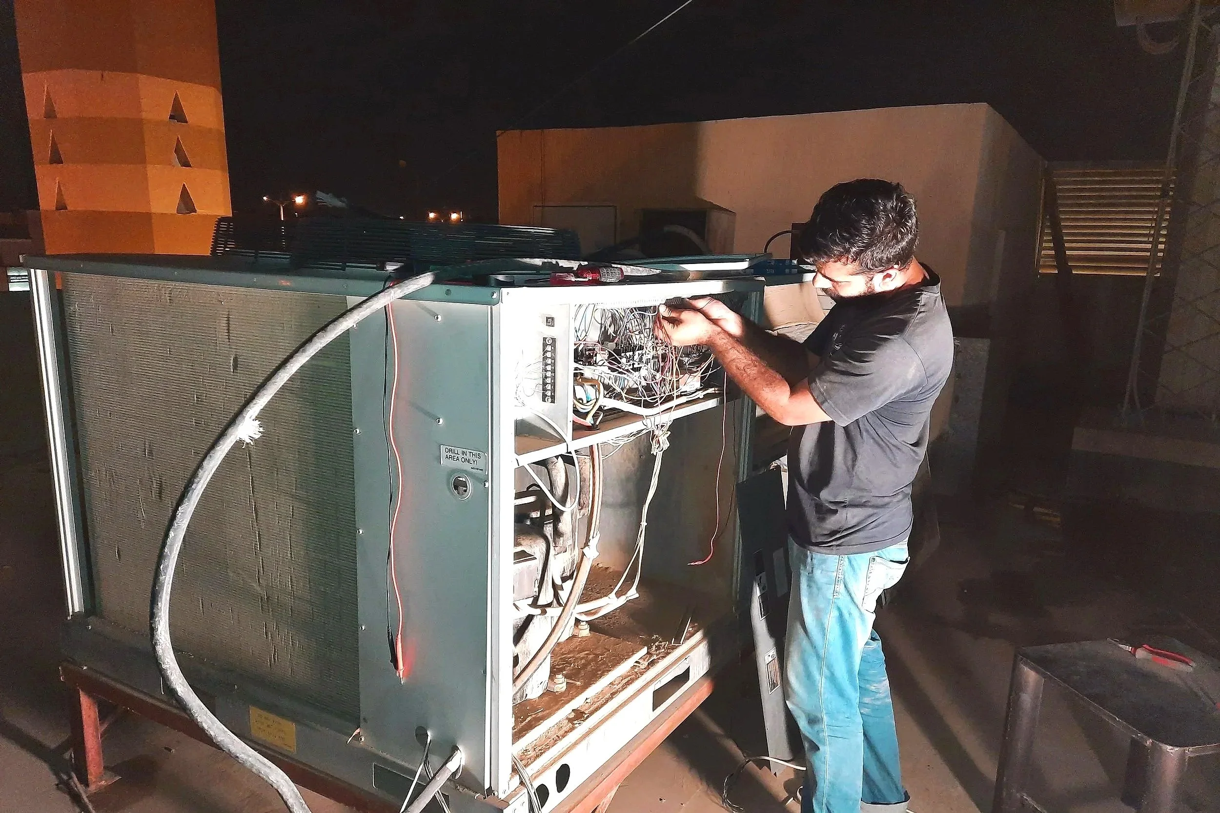 A man repairing or working on an open refrigerator or large appliance outdoors at night. The appliance casing is partly removed, revealing wiring and components inside.