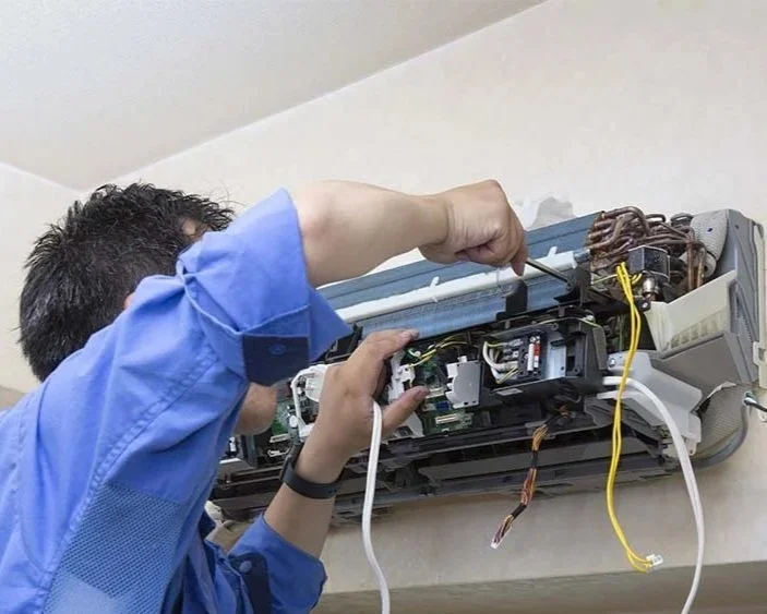 Technician repairing an air conditioning unit mounted on a wall, with the cover removed revealing internal components.