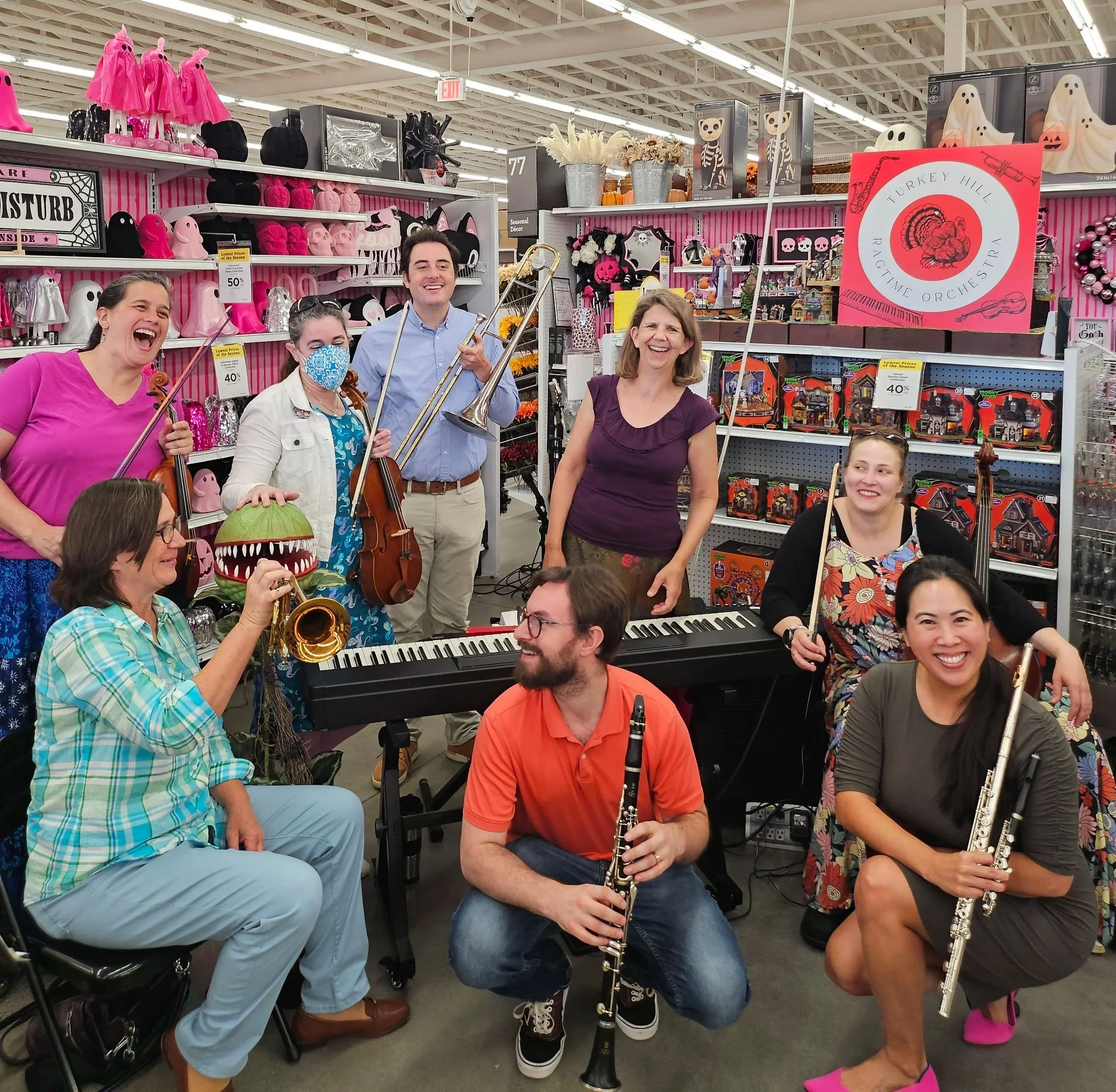 Group of people smiling, playing musical instruments, and enjoying a festive Halloween celebration inside a store aisle decorated with Halloween-themed decorations such as ghosts, skulls, pumpkins, and spooky signs.