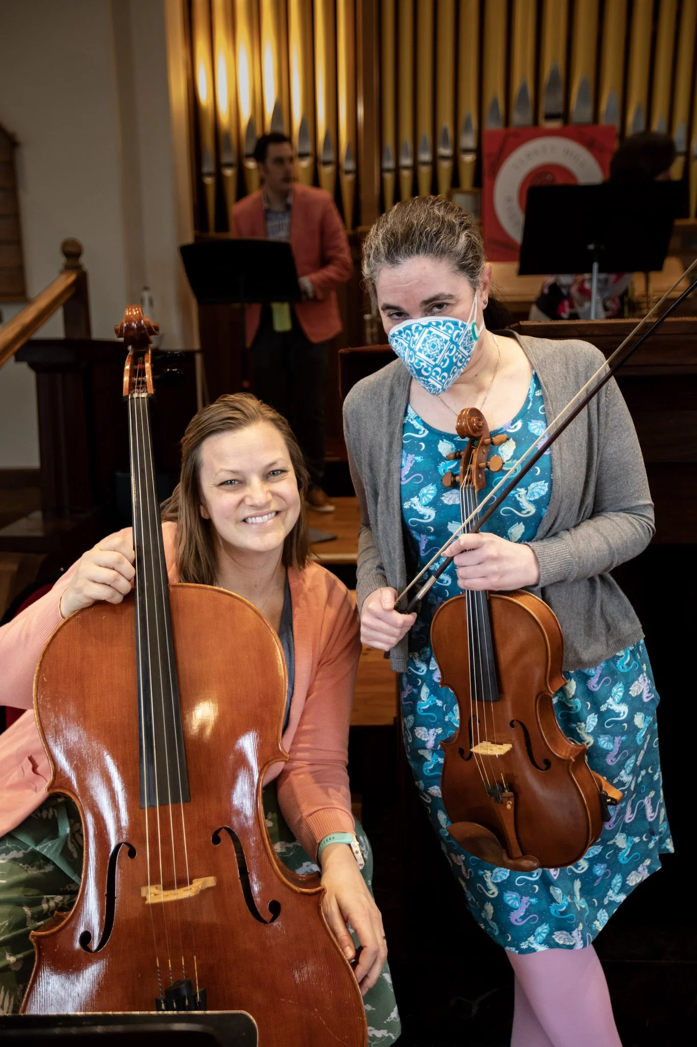 Two women holding violins indoors, one smiling at the camera and the other wearing a blue patterned face mask, with a man in a salmon blazer and a music stand in the background.