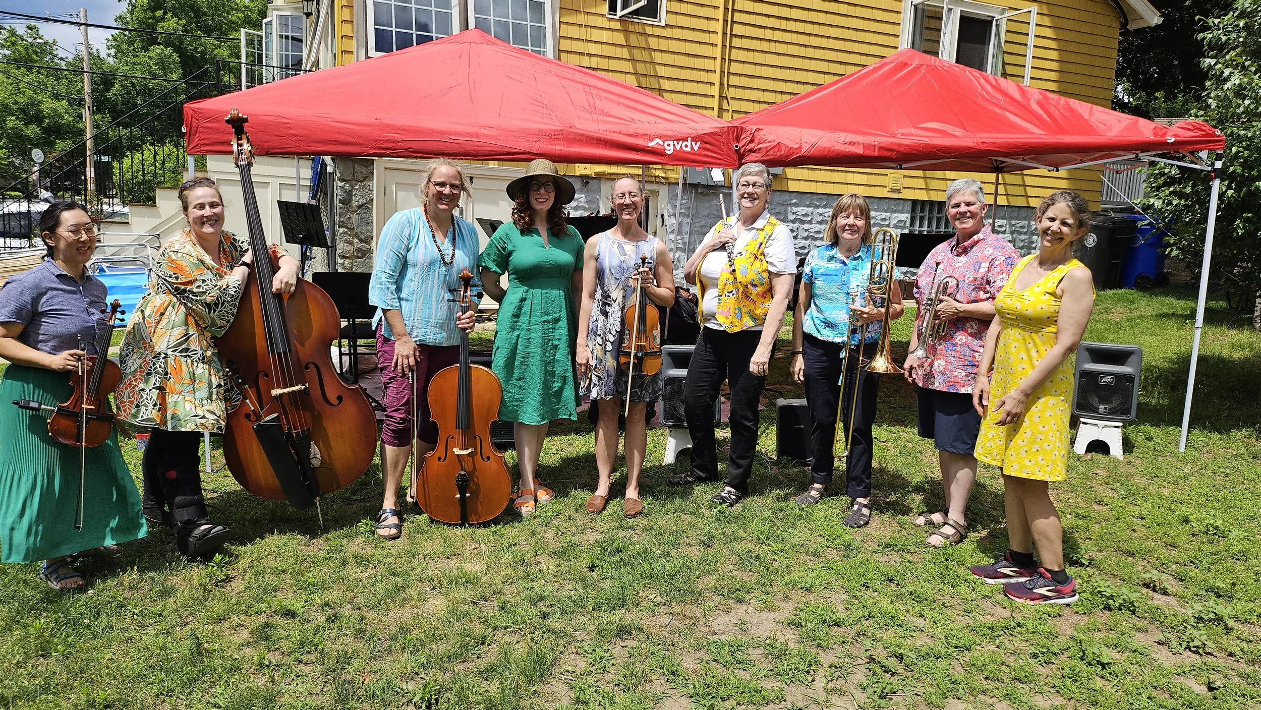 A group of women in colorful summer dresses holding musical instruments, standing under a red canopy outdoors in front of a yellow house with an open window, green grass, and trees.
