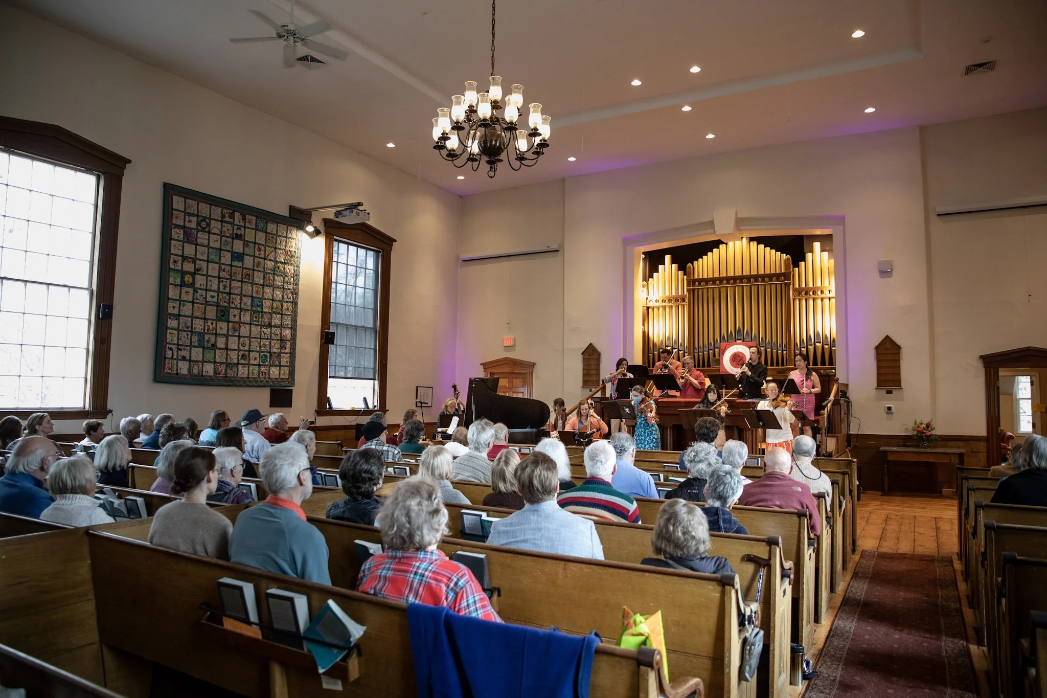 A large group of people attending a concert inside a church or concert hall. The audience is seated on wooden pews or benches, facing forward. On the stage, musicians are performing with some standing and some seated, playing instruments including a 