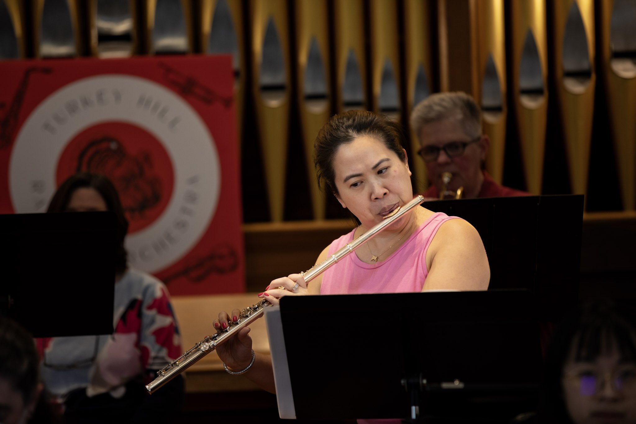 Woman in pink sleeveless top playing a flute during a music performance with musician in the background and a red banner with a white circle and black text.