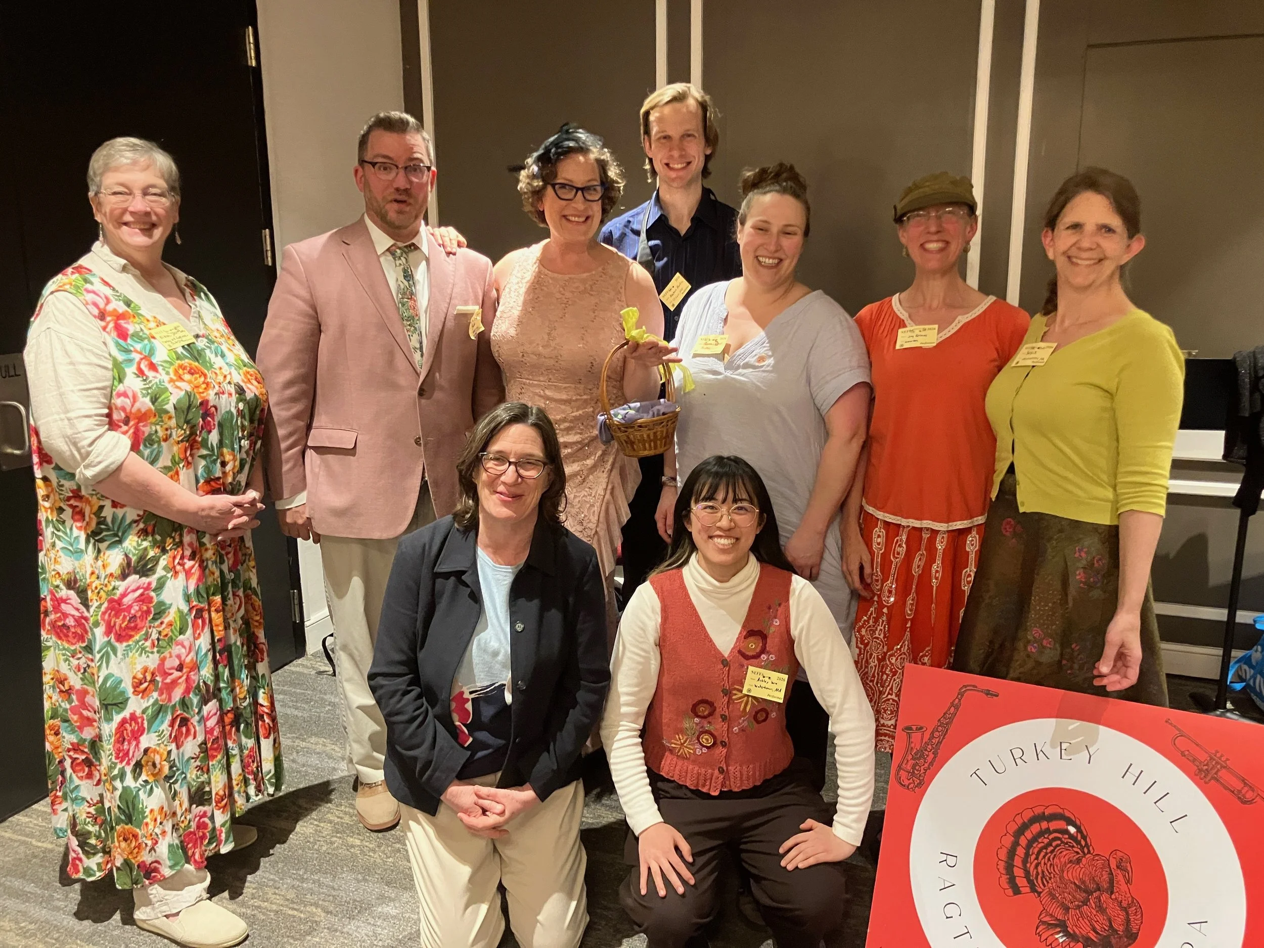 Group of eight smiling people at a social event, standing in front of beige and dark walls. Some are wearing colorful, patterned clothing, and a few have name tags. One person is holding a small basket with a green ribbon. There is a red and white ba