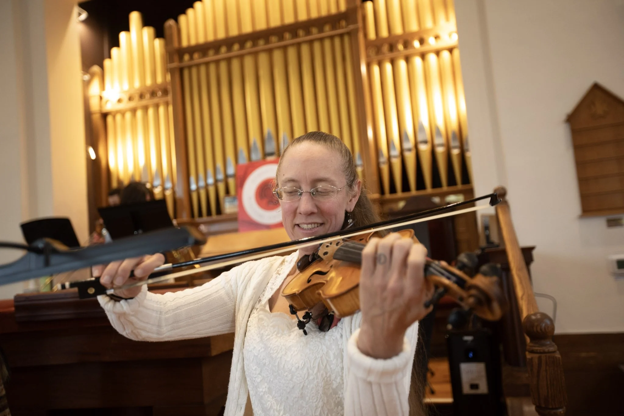 A woman in a white sweater playing a violin in a room with wooden pipes in the background.