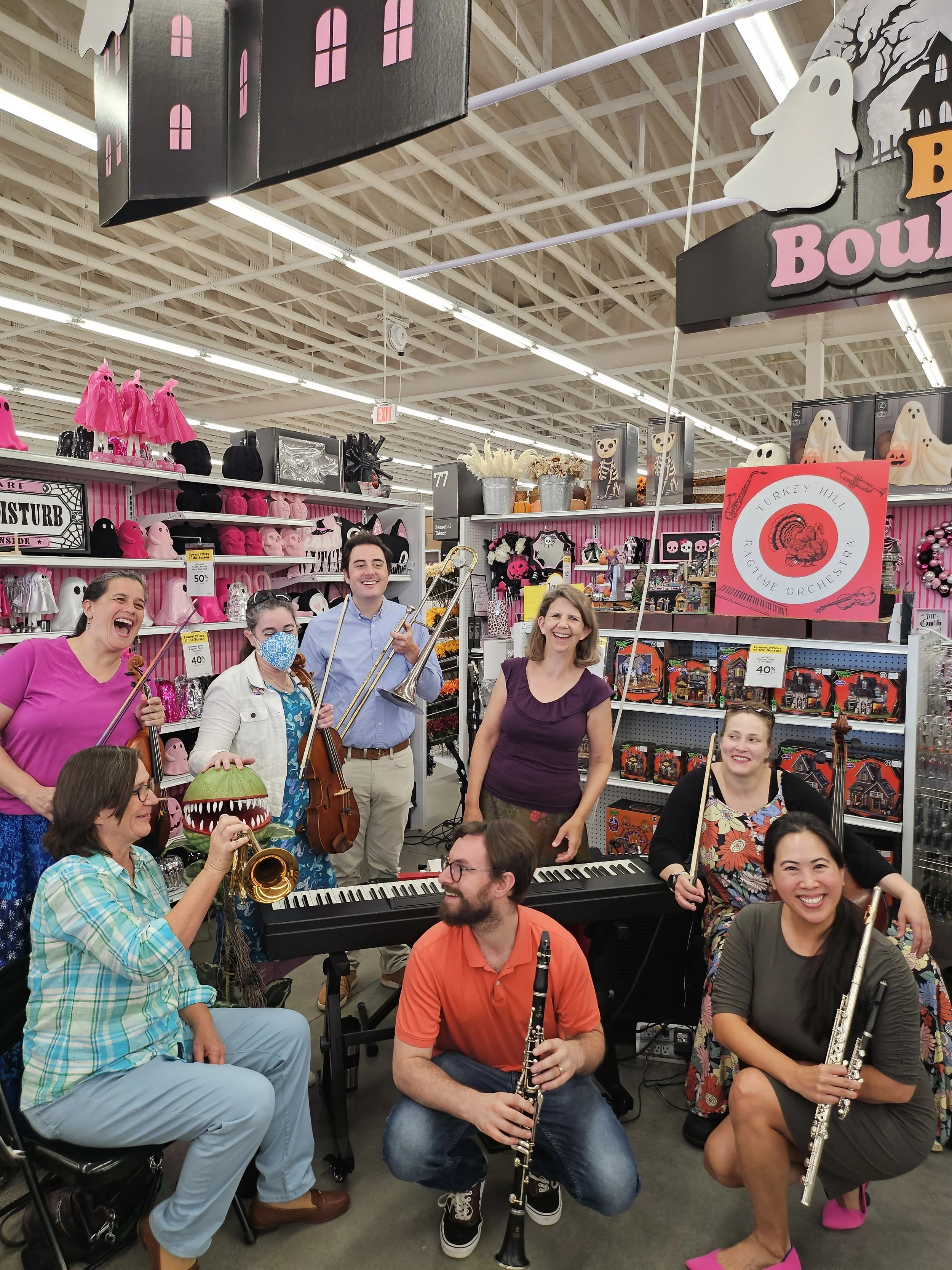 Group of people playing musical instruments and smiling inside a retail store decorated for Halloween with ghost and pumpkin decorations.