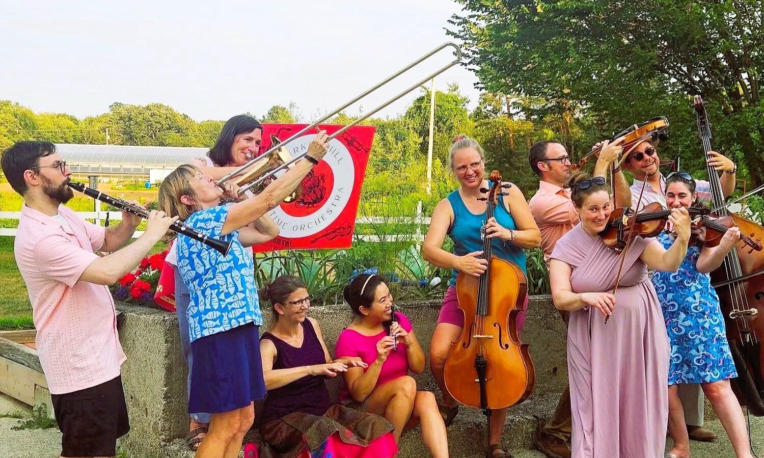 A group of musicians playing string and wind instruments outdoors, smiling and enjoying a performance in a garden with trees and plants.