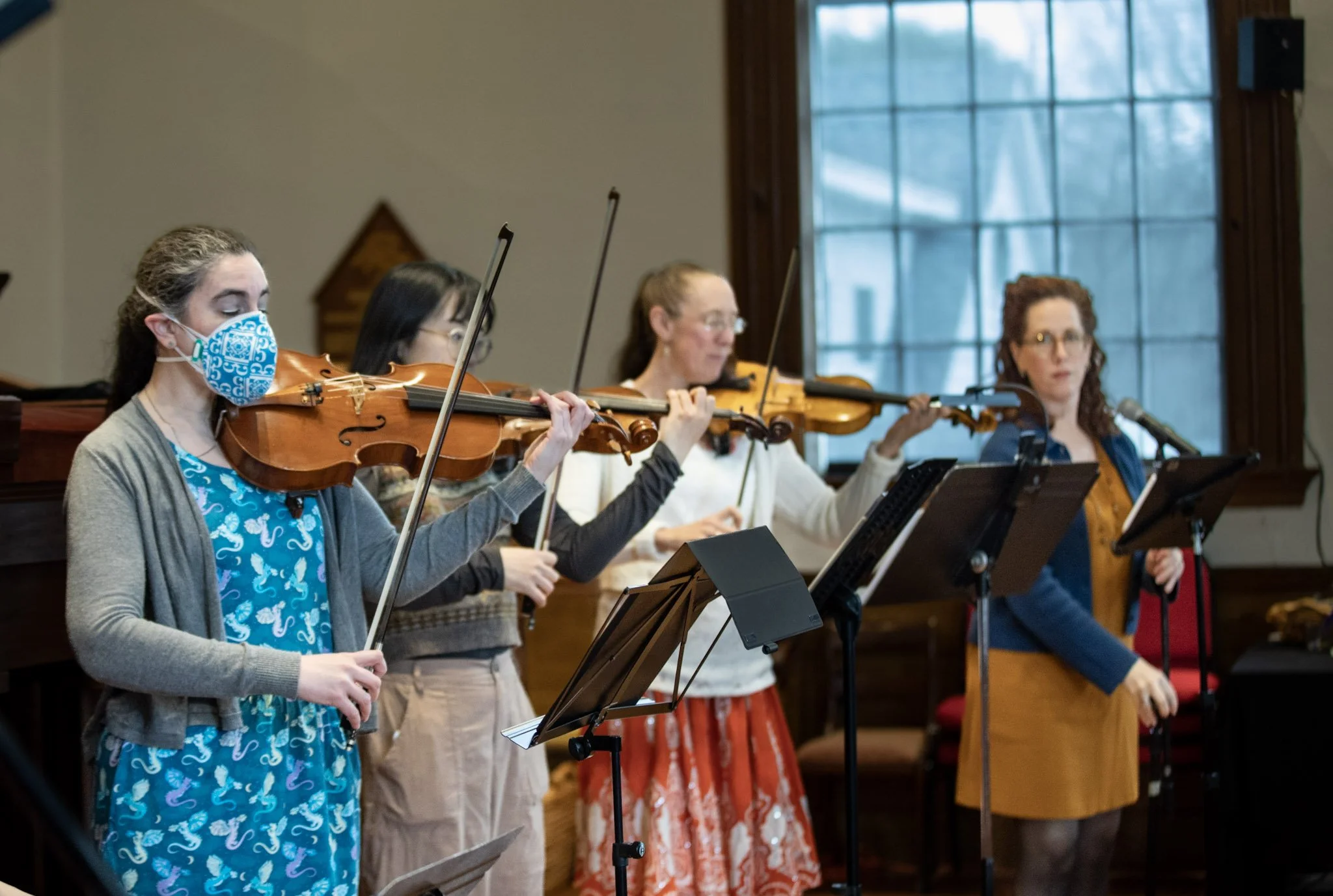 Four women are playing violins during a rehearsal or performance in a room with large windows.