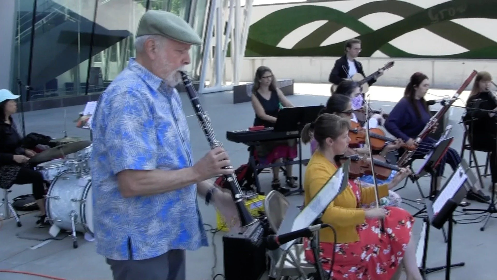 An outdoor musical performance featuring a group of musicians, including an older man playing a clarinet in the foreground and several women playing instruments such as violin, guitar, drums, and keyboard in the background, with a modern building and