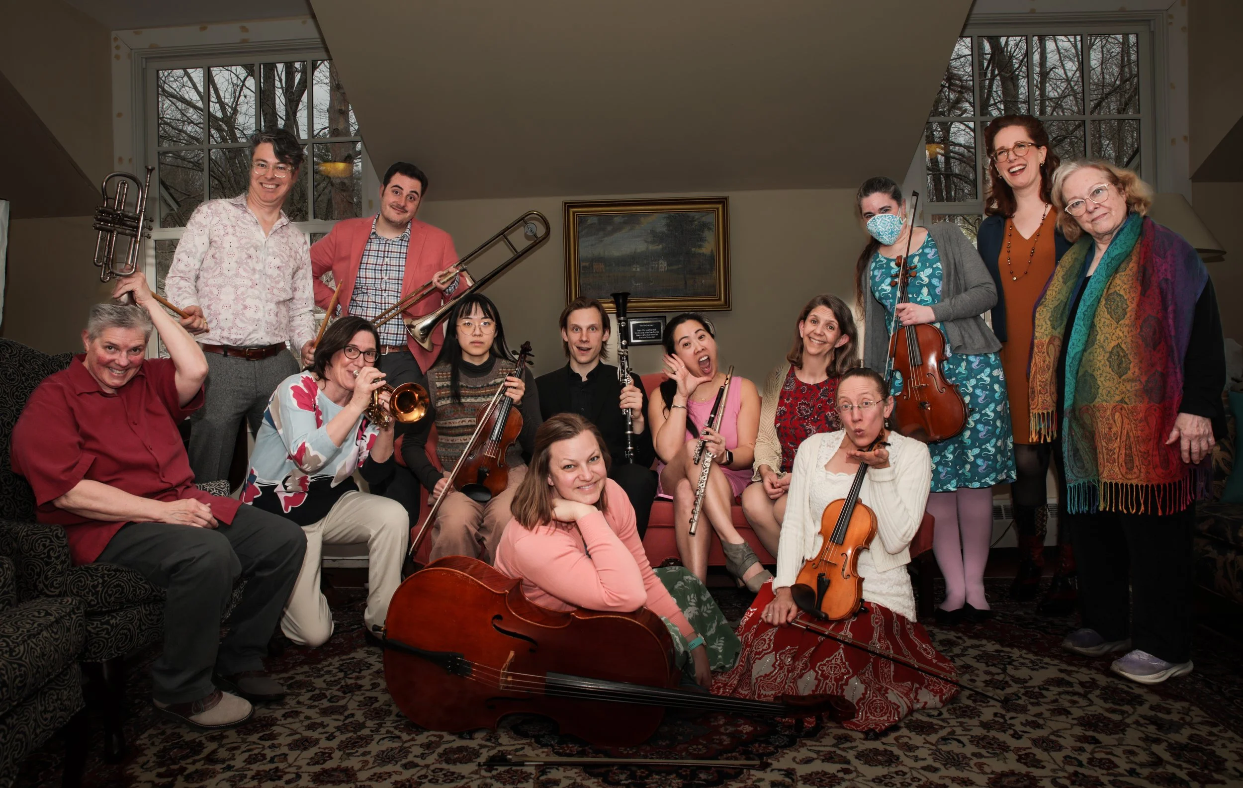 A group of people posing for a photo in a living room, holding musical instruments such as violins, a trombone, and a flute, with some wearing masks and colorful clothes.