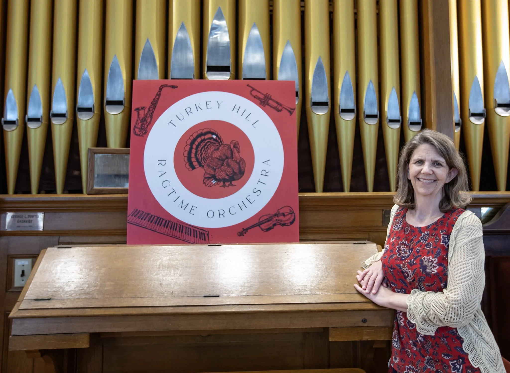 A smiling woman with shoulder-length hair wearing a red and blue patterned dress and a cream-colored cardigan poses with her hands resting on a wooden railing in front of a display of yellow organ pipes. A red circular sign reading 'Turkey Hill Razzm
