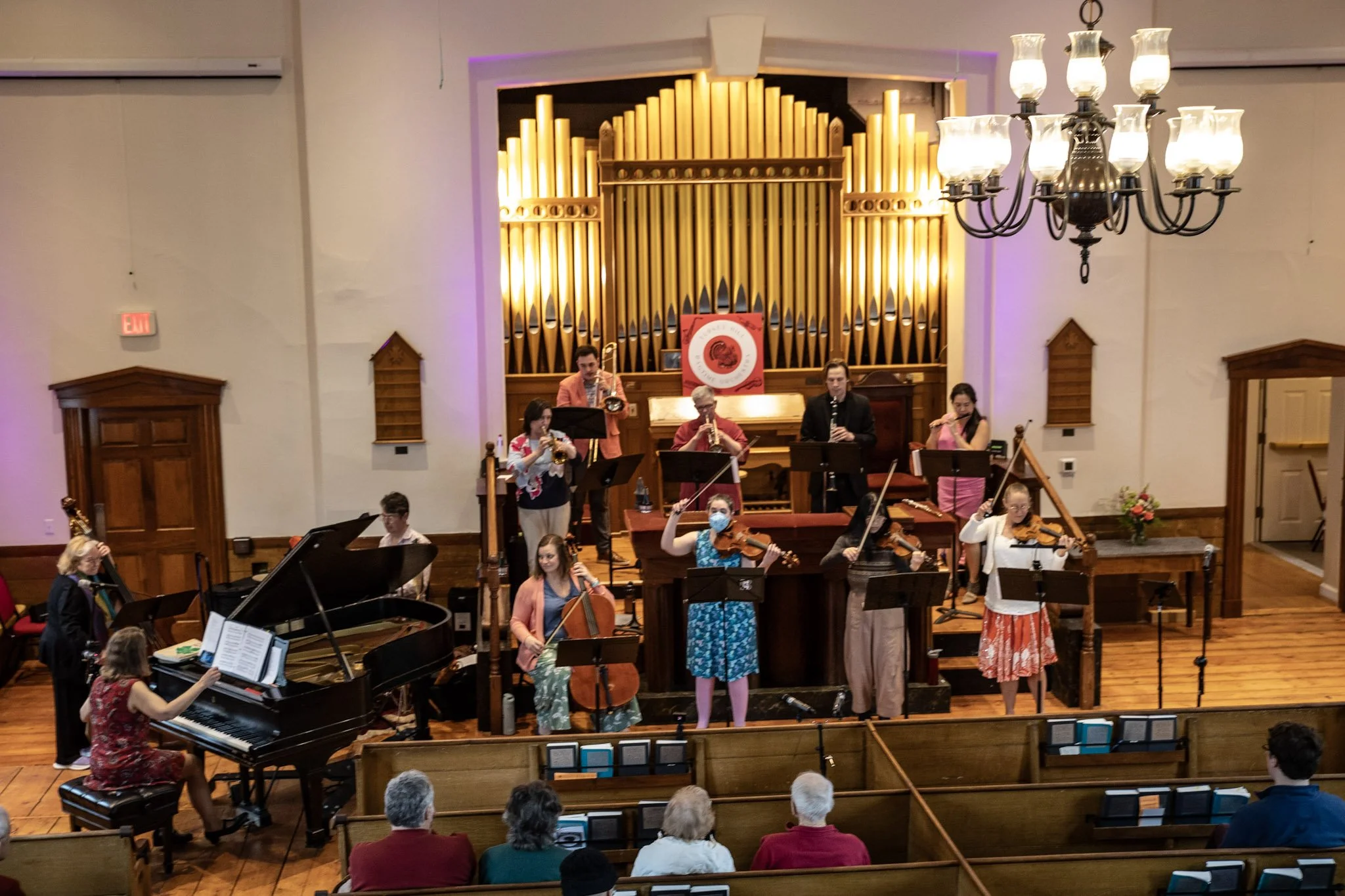 An orchestra performing on stage in a concert hall, with musicians playing various instruments including strings, wind, and piano. The backdrop features large golden organ pipes and a red banner. Audience members seated in front observe the performan