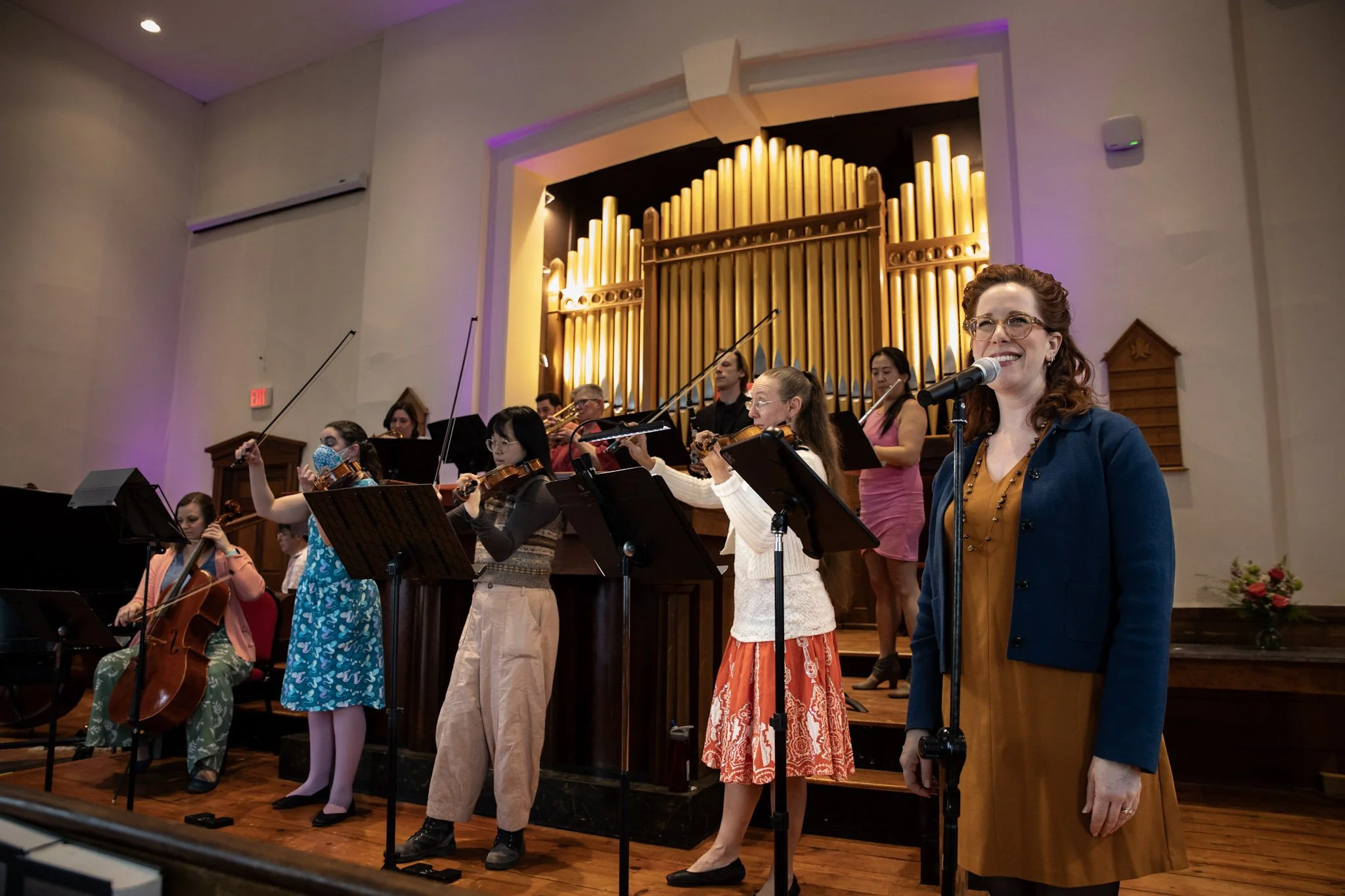 A group of children and women, some holding musical instruments, are performing on stage in a concert hall. The hall features a large pipe organ in the background and warm lighting.