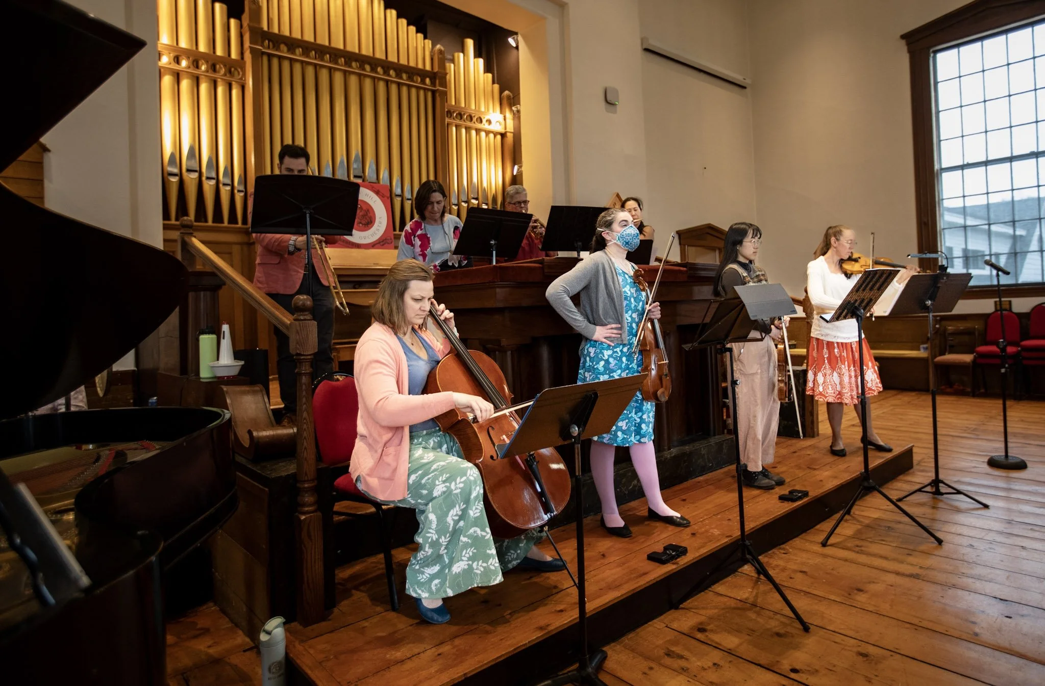 A group of musicians performing on stage in a church, with some playing violins, a cello, and others standing behind music stands, with an organ in the background, some adults and children, one wearing a face mask.