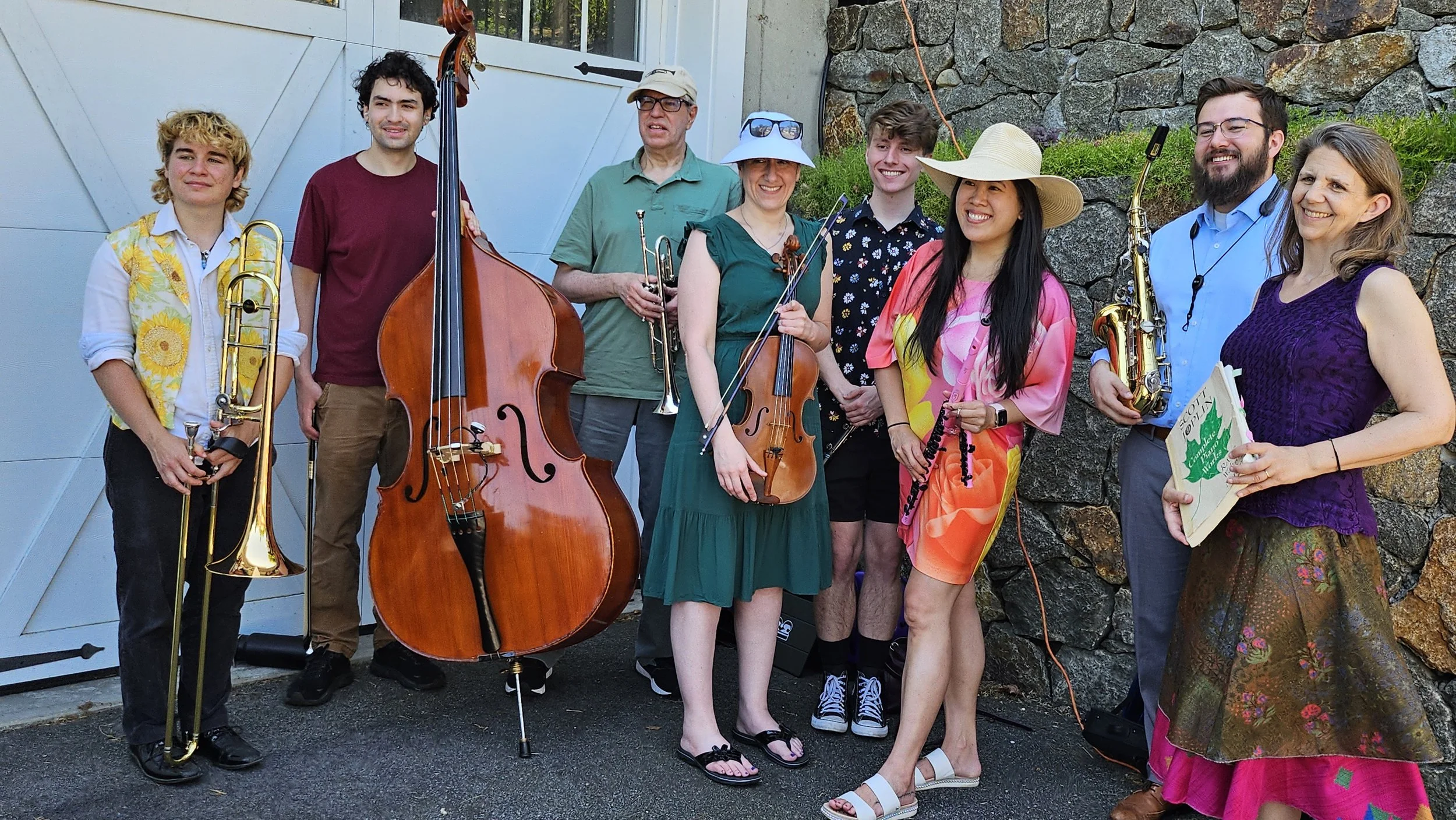 Group of nine people holding musical instruments outside near a rock wall and a house with a garage door.