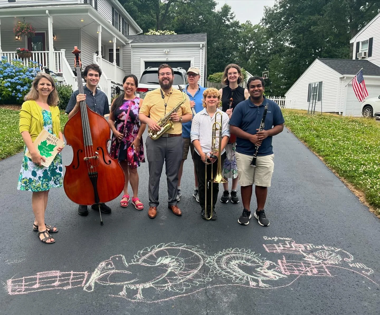 A group of nine people standing on a driveway in front of houses, holding musical instruments, with colorful chalk drawings on the pavement, including a guitar and a dragon.