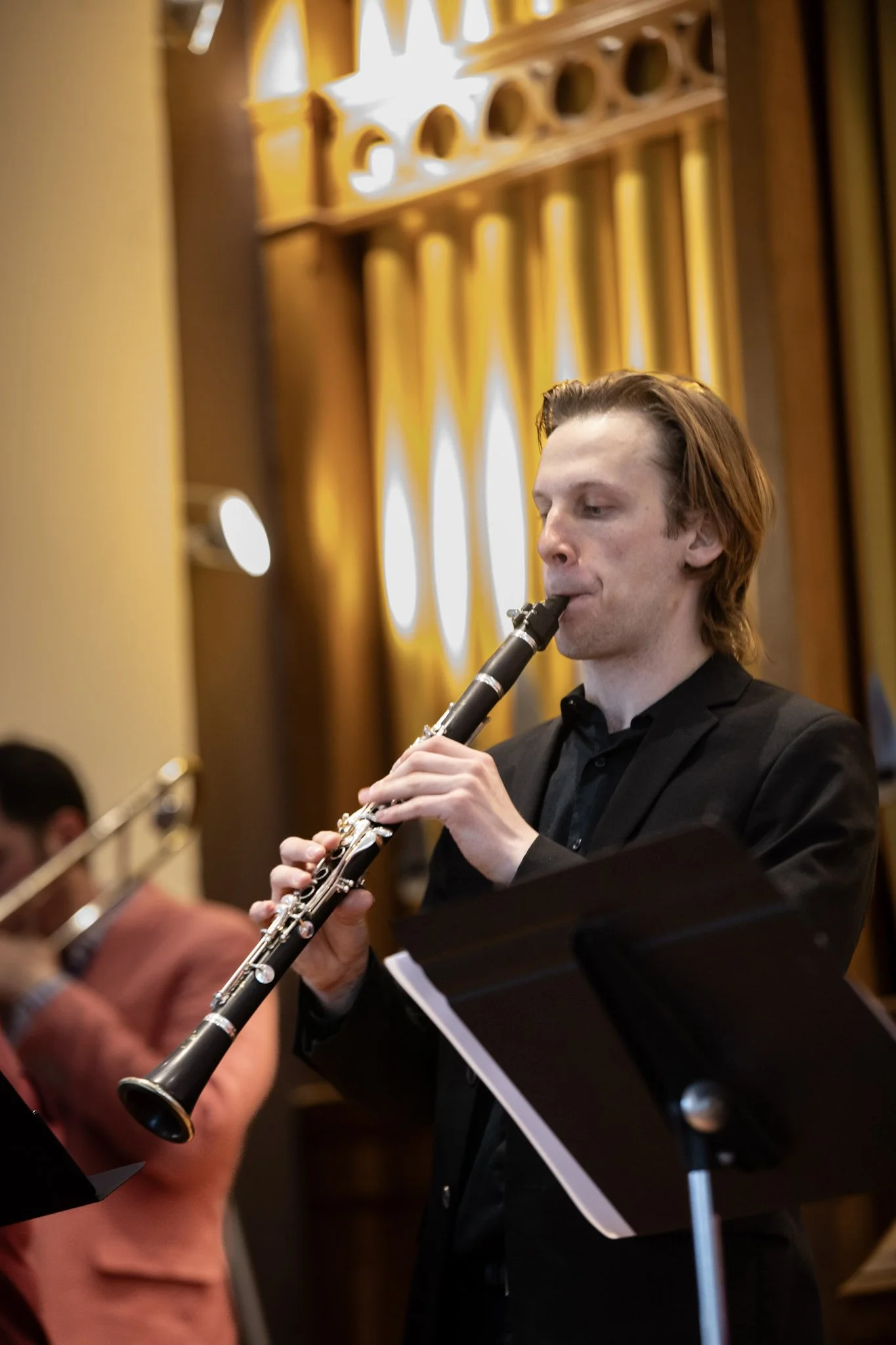 A musician playing a clarinet during a performance, with sheet music on a stand in front of him, in a venue with warm lighting and decorative wood paneling.