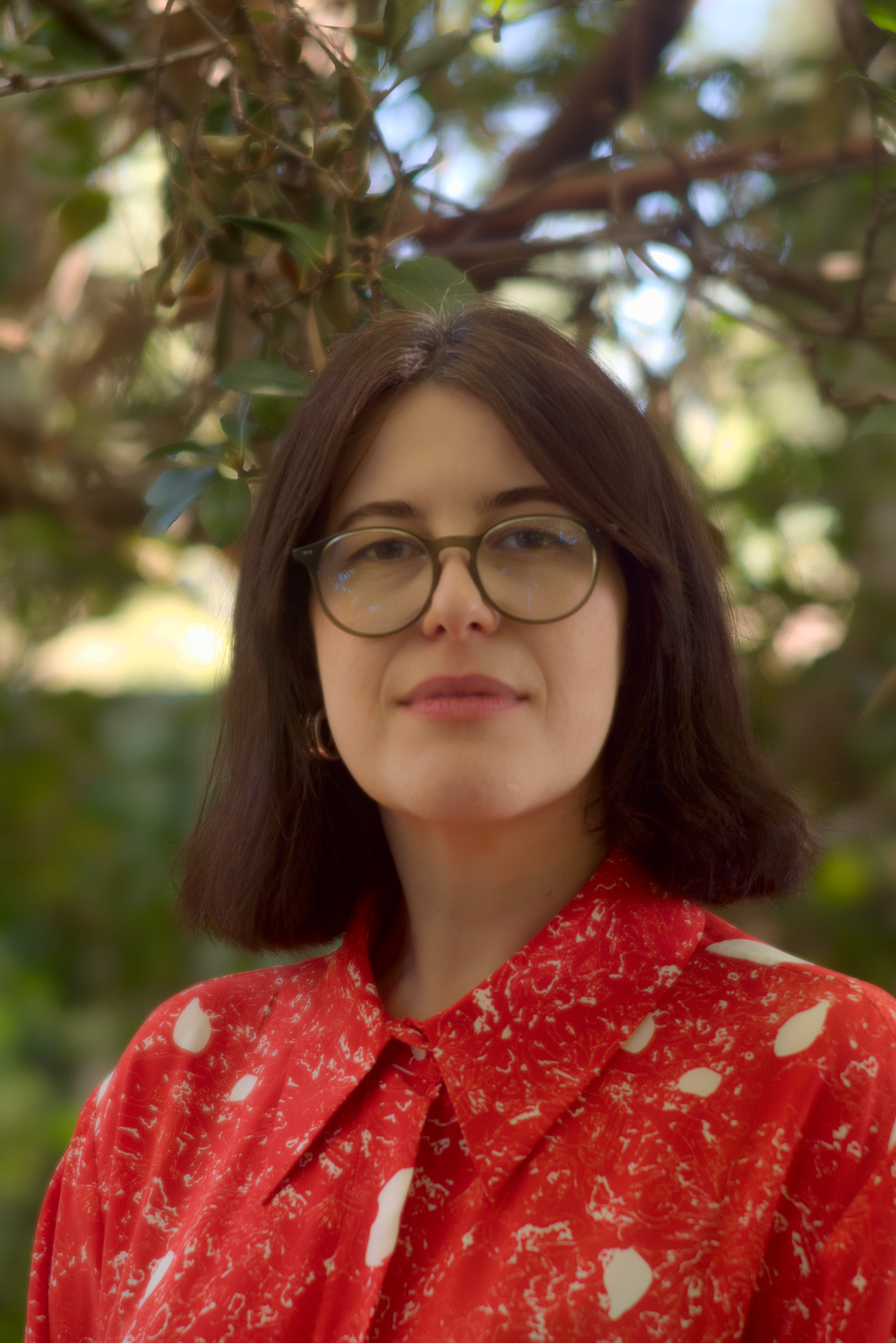 A young woman with shoulder-length brown hair and glasses, wearing a red patterned top, standing outdoors amid green foliage and branches.