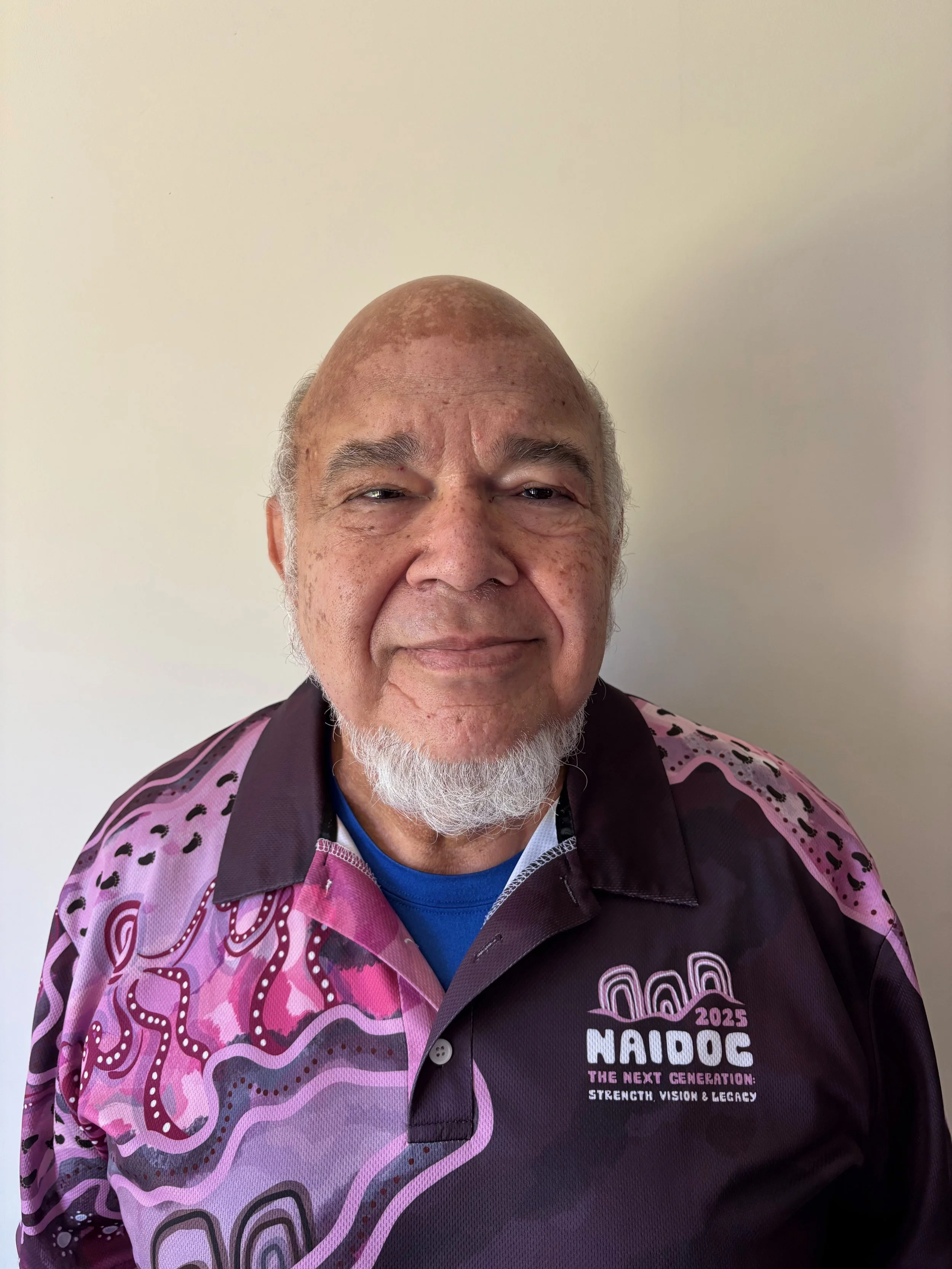 A man smiling wearing a pink and purple shirt with 'NAIDOC 2023' logo and text that reads 'NAIDOC THE NEXT GENERATION: STRENGTH, VISION & LEGACY' against a plain white wall.