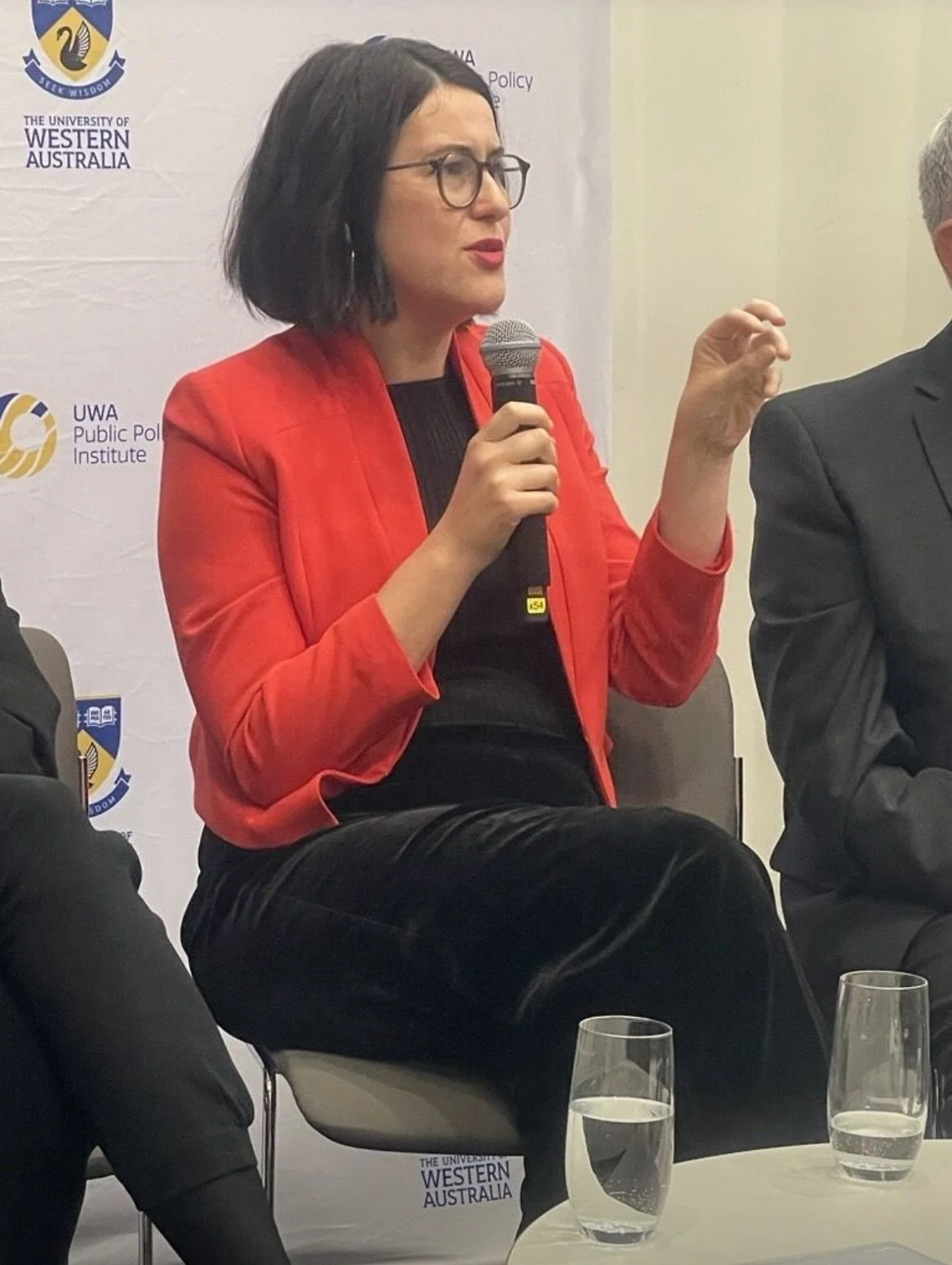 A woman with black hair, glasses, and wearing a red blazer, speaking into a microphone during a panel discussion at the University of Western Australia, with a banner displaying the university's logo in the background.