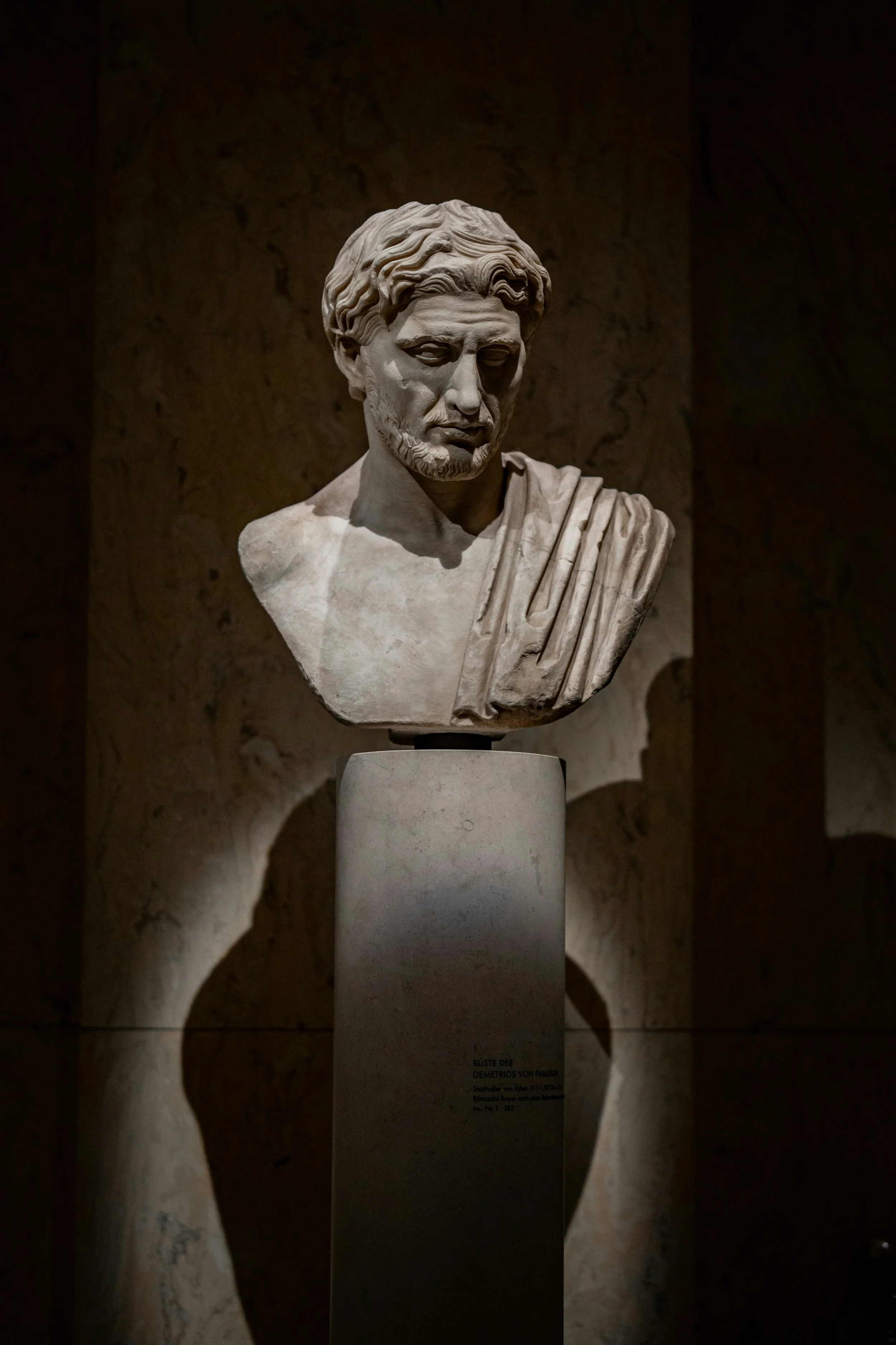 Marble bust of an ancient Greek man with wavy hair and beard, displayed on a pedestal in a dimly lit museum setting.