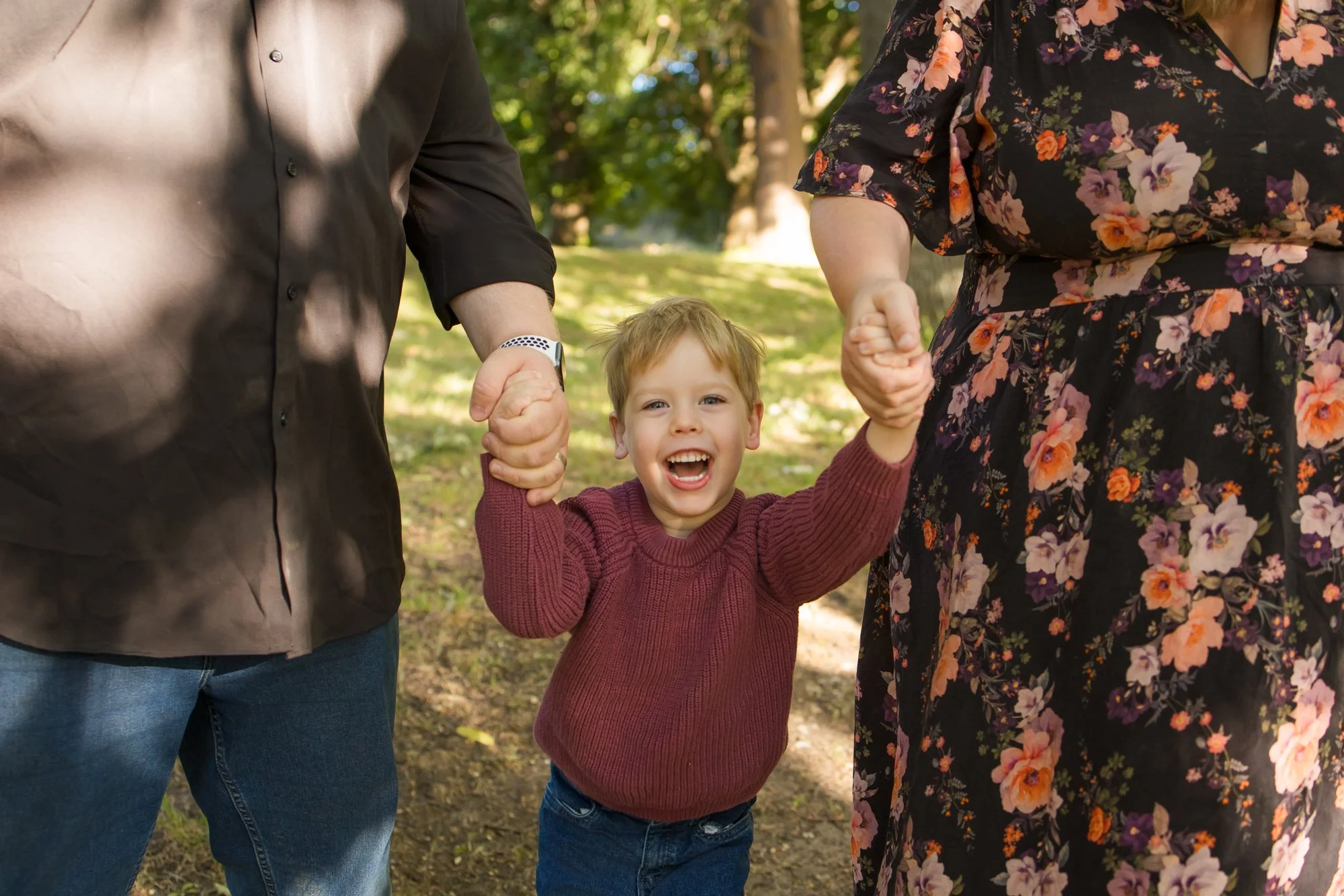A young boy with blond hair, smiling and holding hands with his parents, in an outdoor park with trees and greenery for a family photography session in Melton West