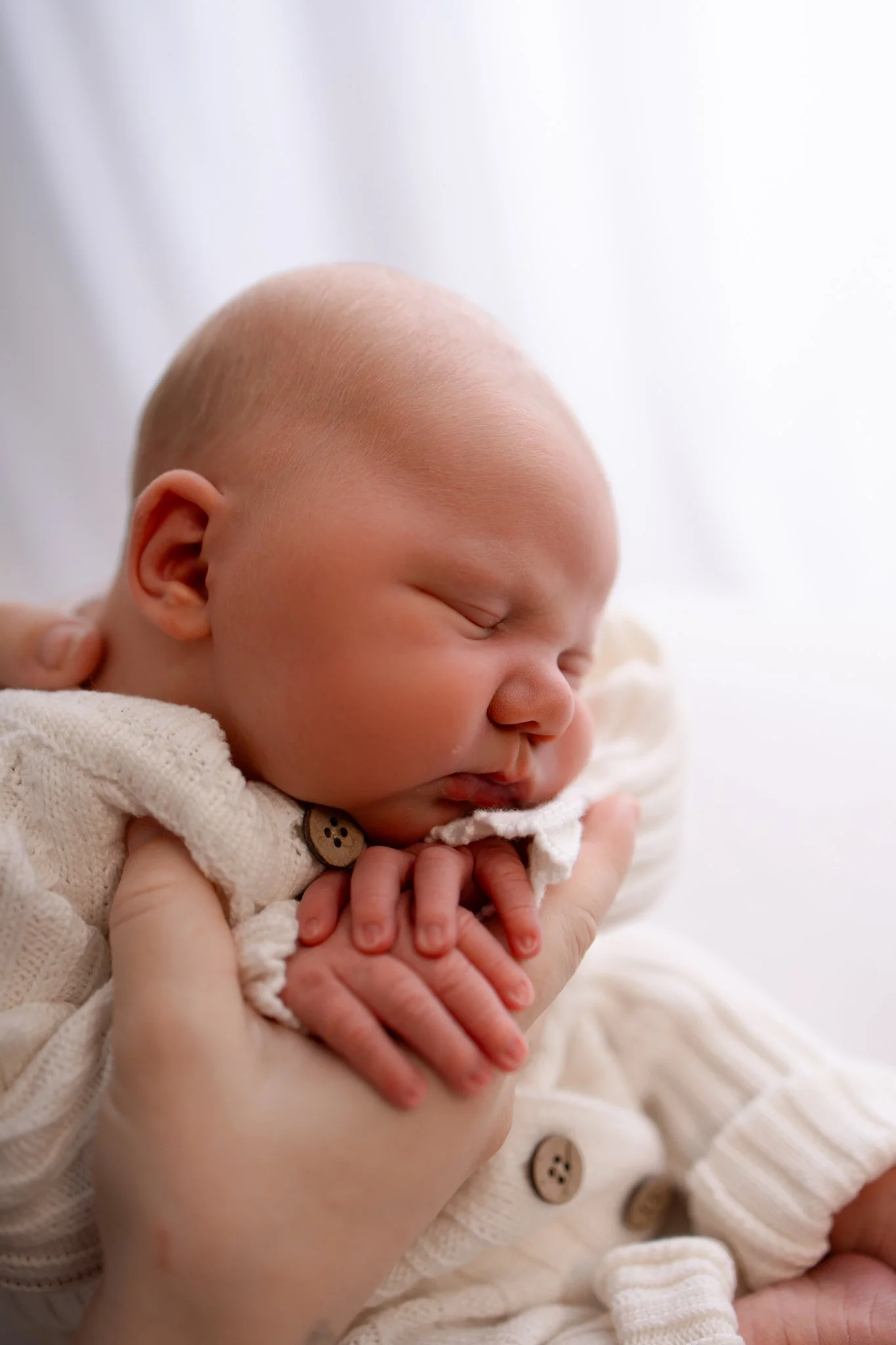 Close-up of a sleeping baby with a smooth head, cradled in someone's hands, wearing a cream-colored knit garment with buttons.