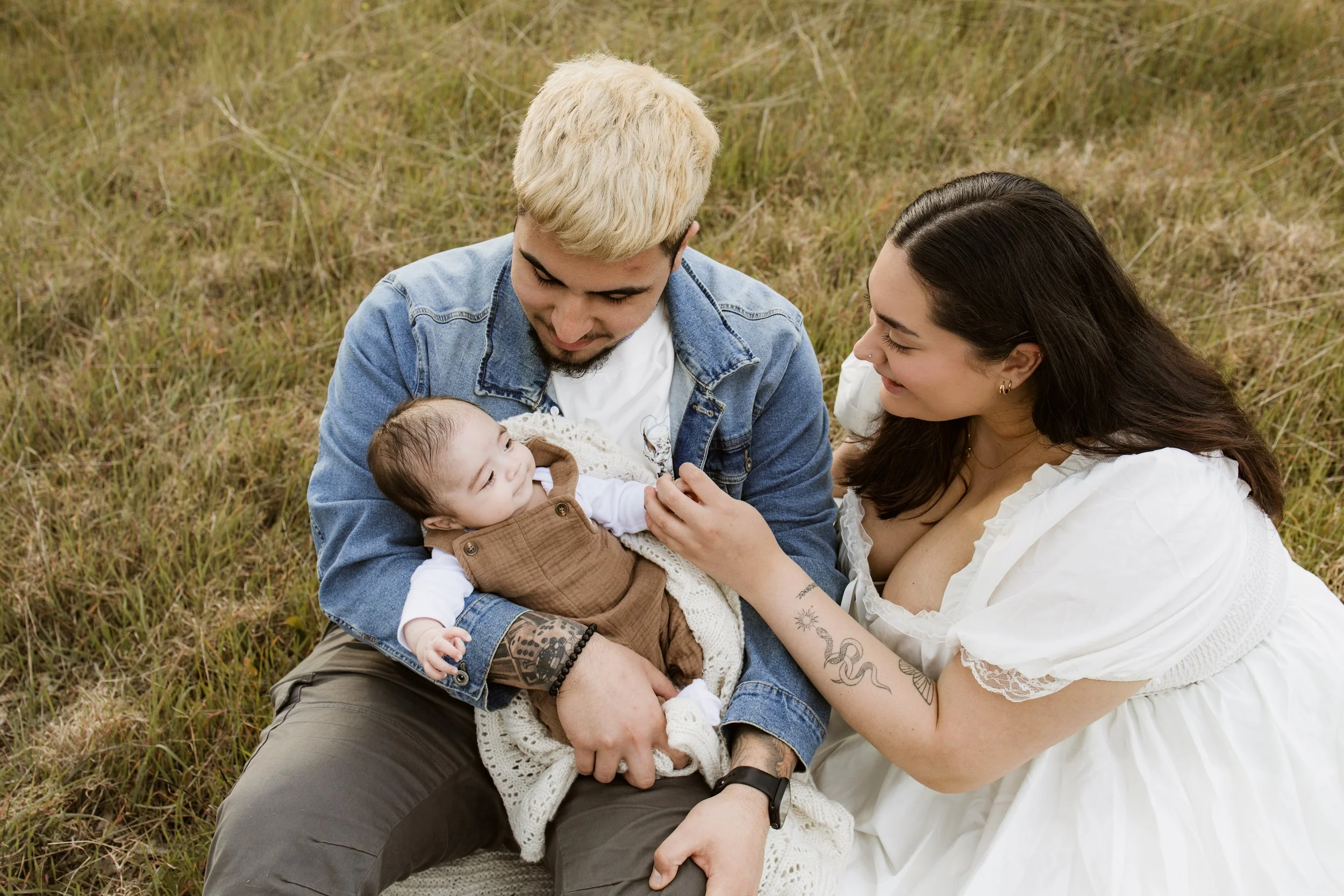 A dad holding a baby while mum is in a white dress looking at them, all sitting on grass in an outdoor setting for their family photos.
