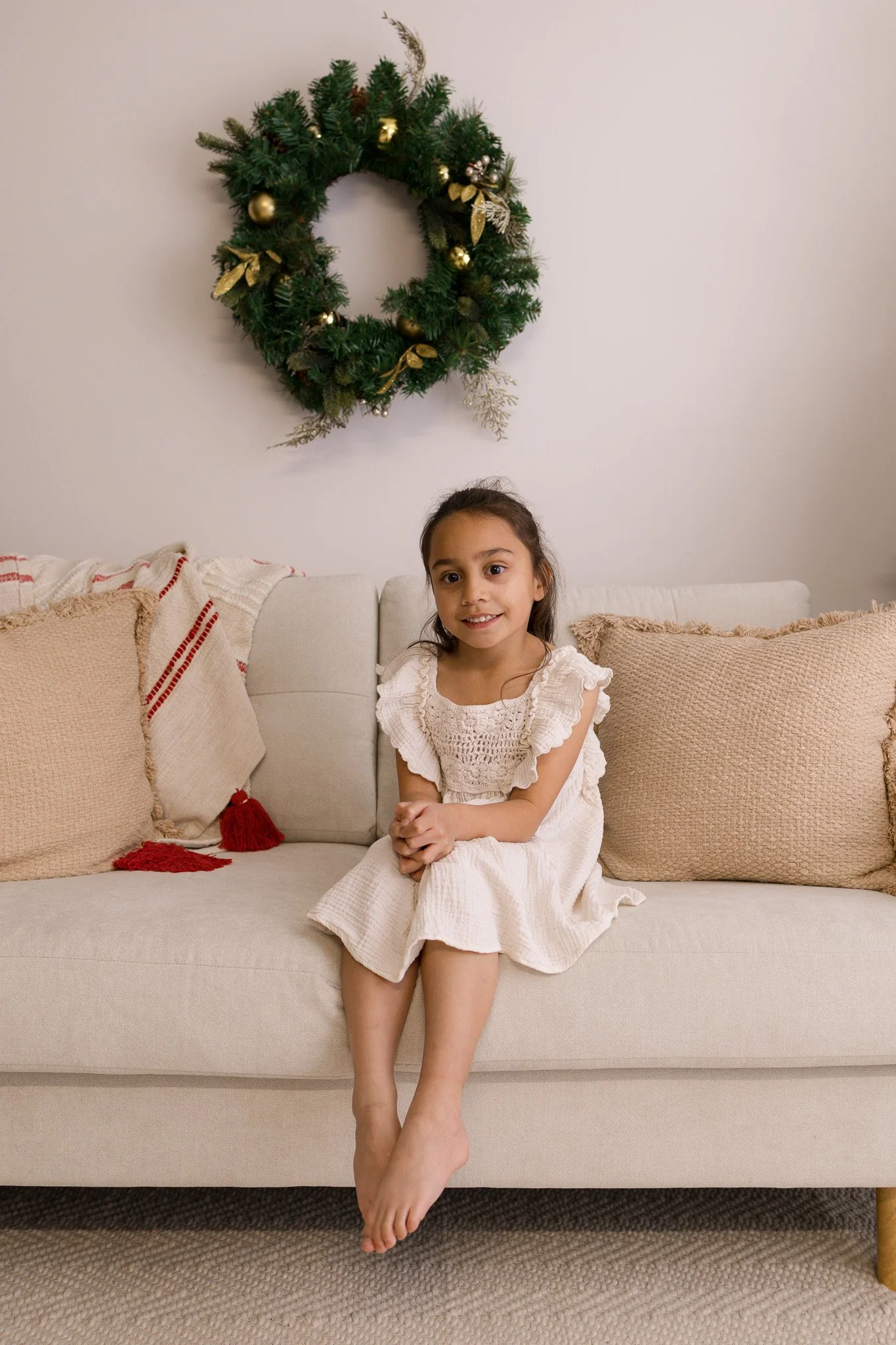 A young girl in a white dress sitting on a beige sofa with a Christmas wreath hanging on the wall behind her. Christmas Mini's Melbourne