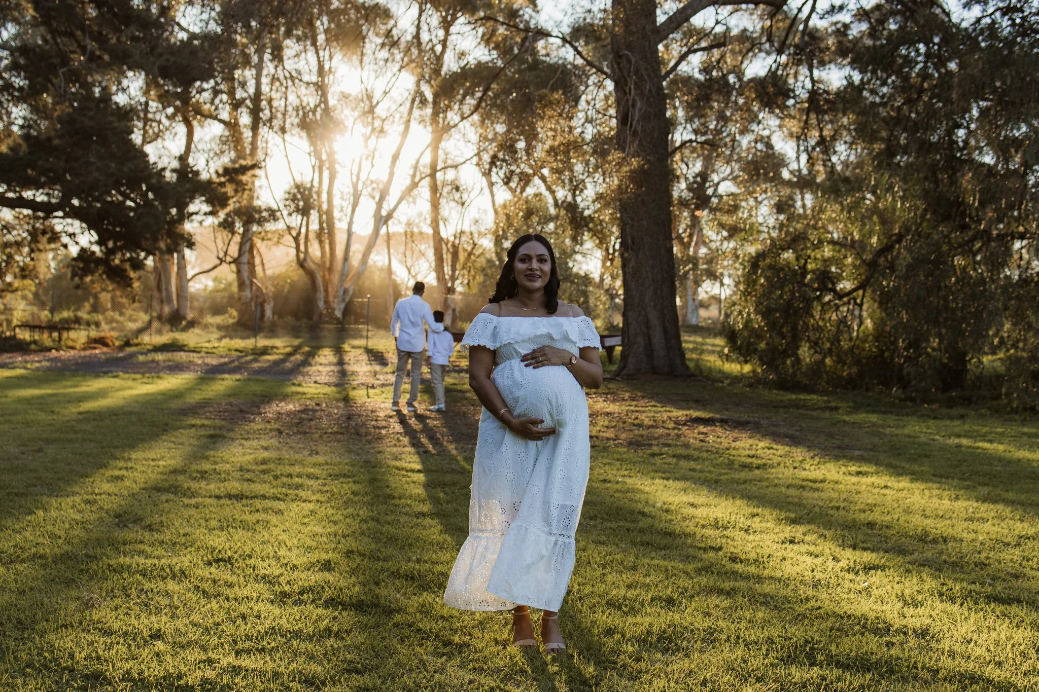 Maternity photography session with mum looking at the camera and her son and husband in the background looking into the sunset