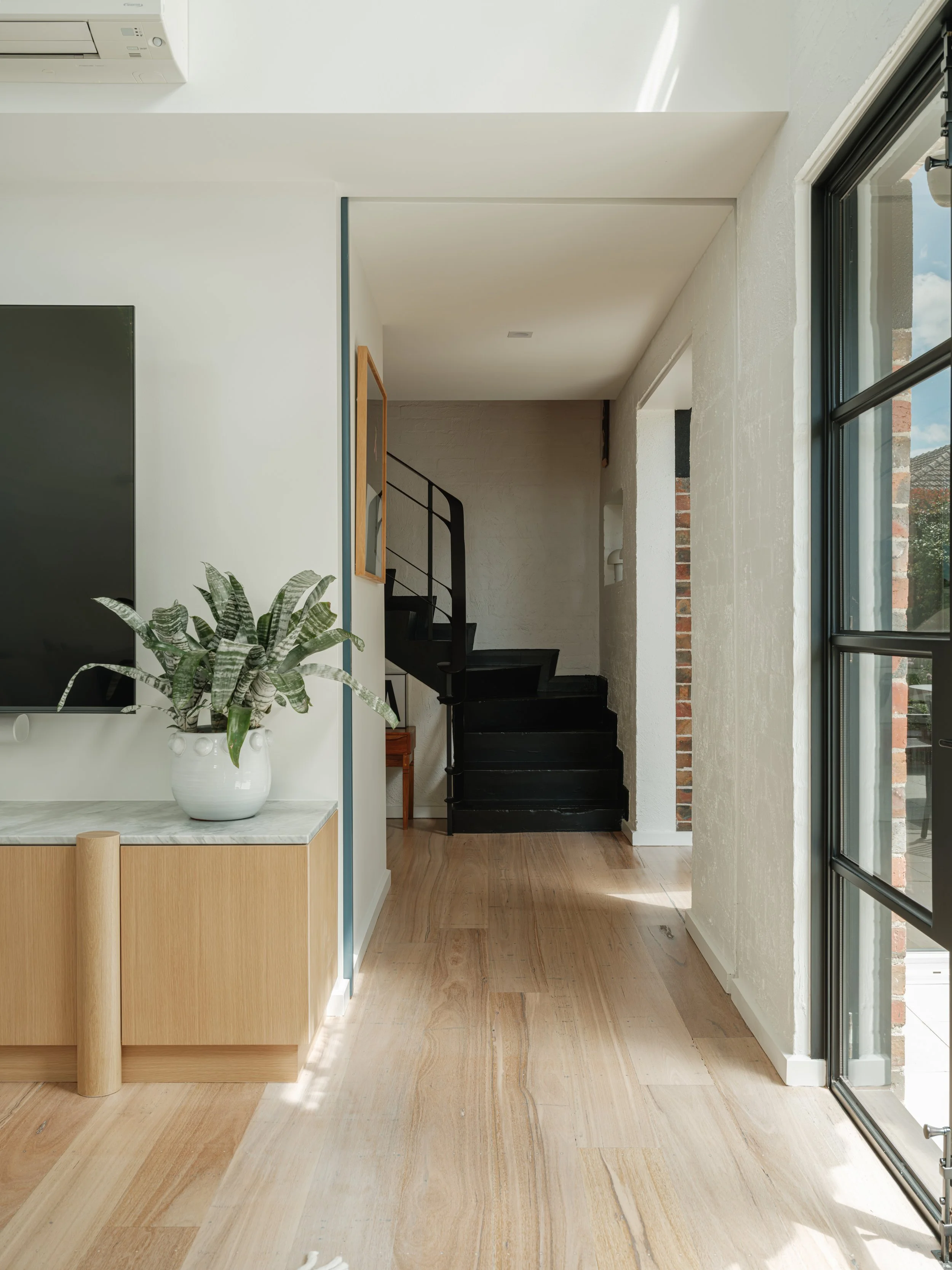 Stinton Project — modern interior space with wooden floors, a plant in a white pot on a light wood cabinet, black staircase railing, and large windows with black framing letting in natural light, Kingston Built, Geelong
