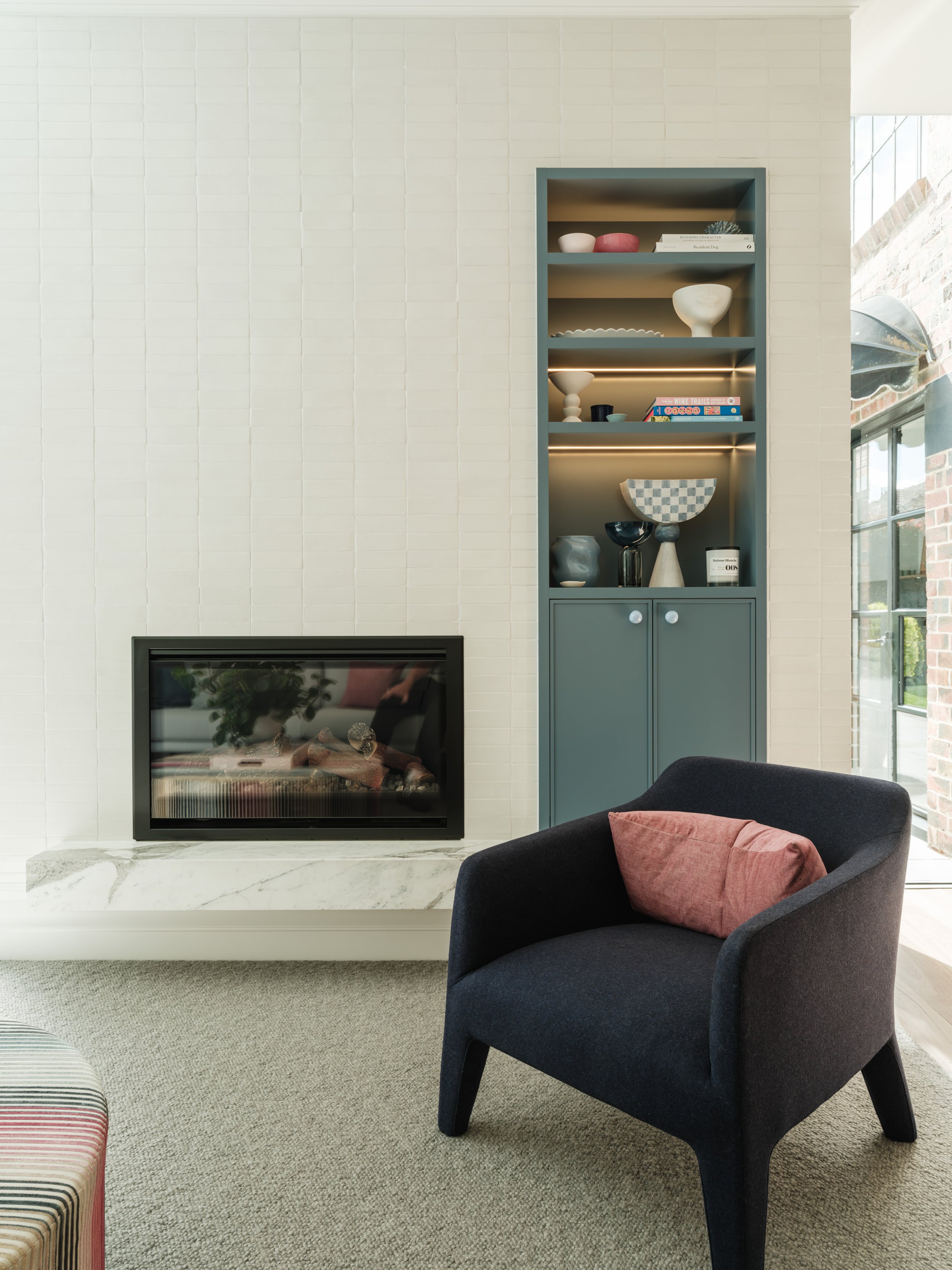 Stinton Project — modern living room with a black armchair, a fireplace with a white marble mantel, and a blue cabinet filled with decorative objects. Large windows allow natural light into the space, Kingston Built, Geelong