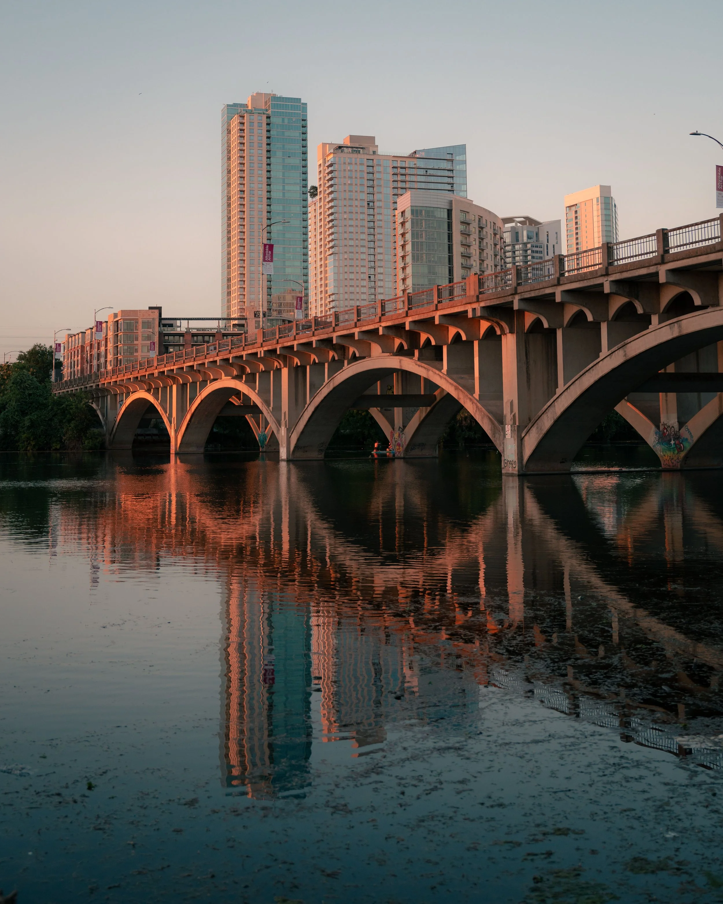 Reflections of modern high-rise buildings and a bridge over a river at sunset.
