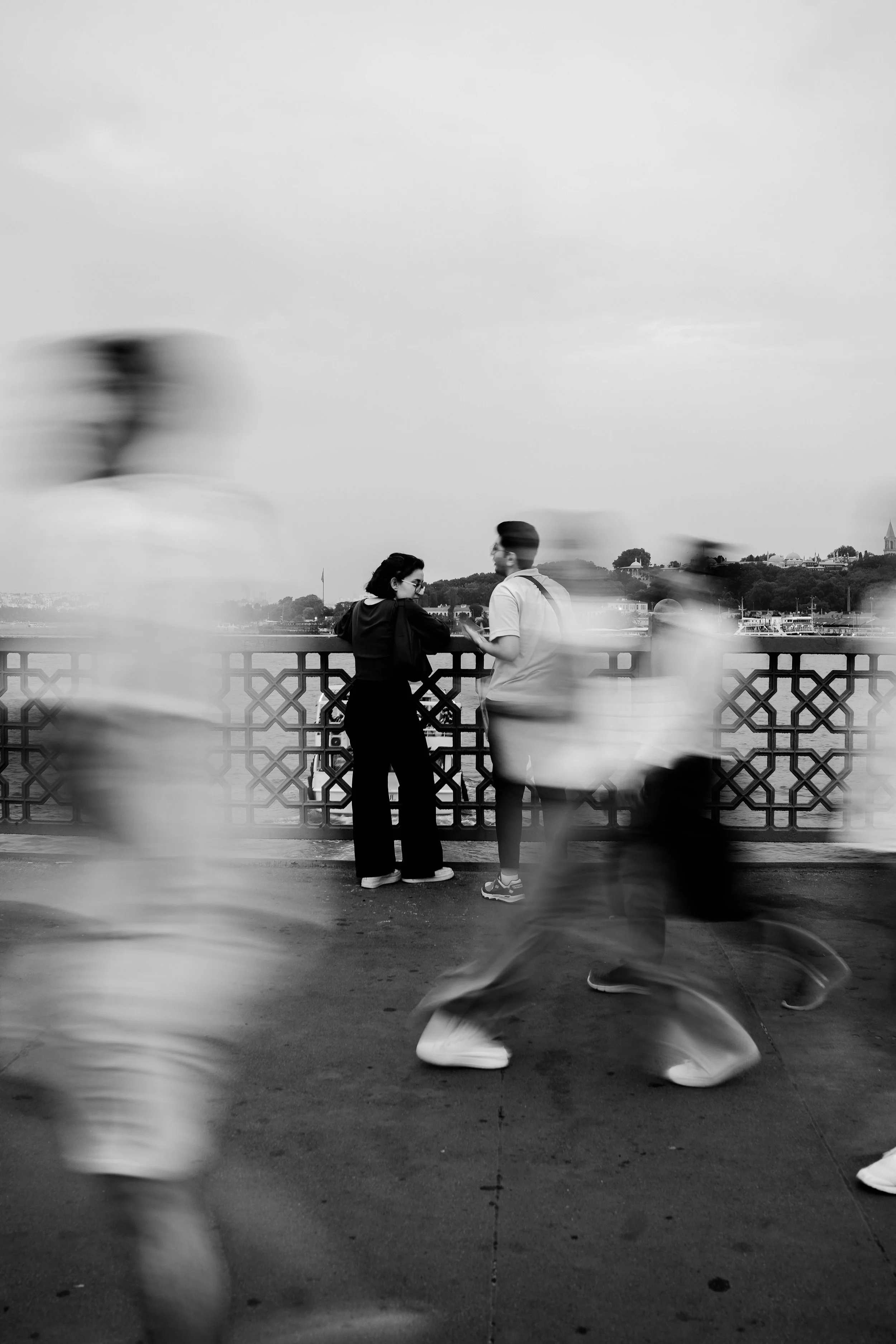 Two people standing by a railing on a riverside pathway, one woman and one man, engaged in conversation. Blurred figures and blurred pedestrians walk past in the foreground, while buildings and a hill are visible in the background.