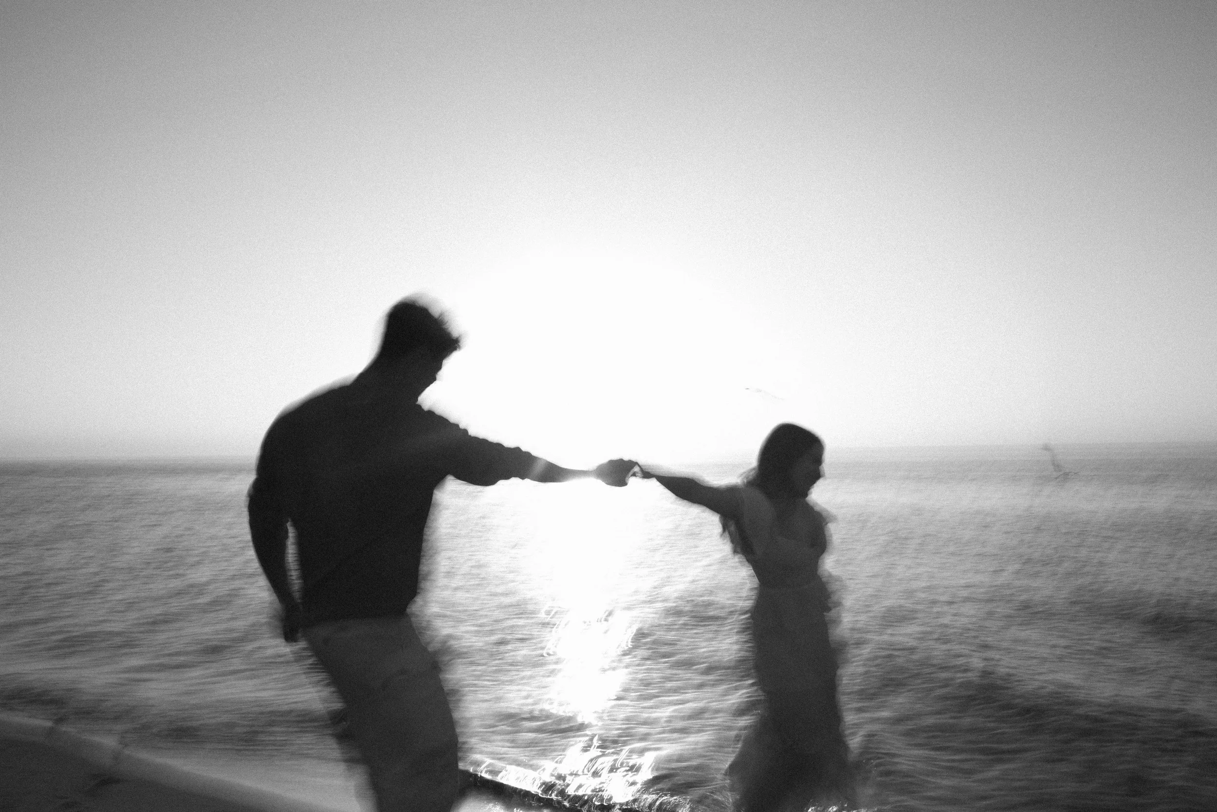 Black and white photograph of a man and girl running hand in hand along a beach at sunset, with waves and ocean in the background.