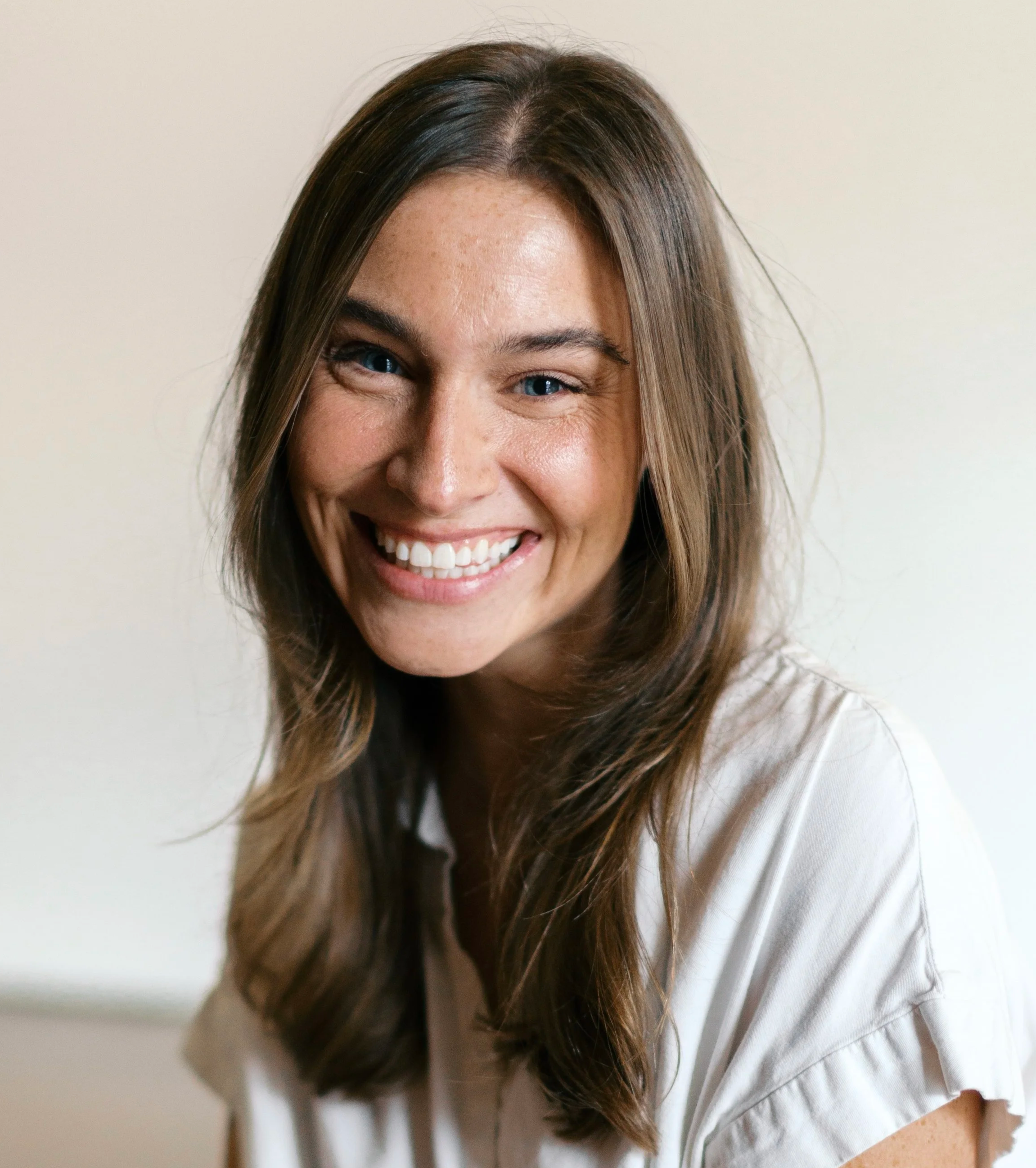 Close-up of a smiling woman with long brown hair and light skin, wearing a white top, against a plain light background.