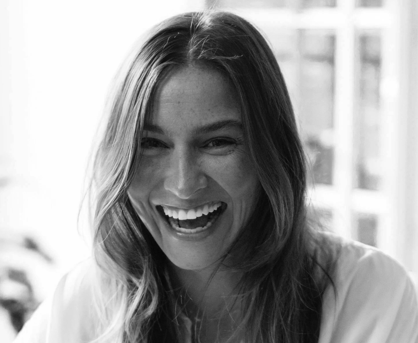 A woman with long hair laughing brightly, showing her teeth, in a black and white photo.
