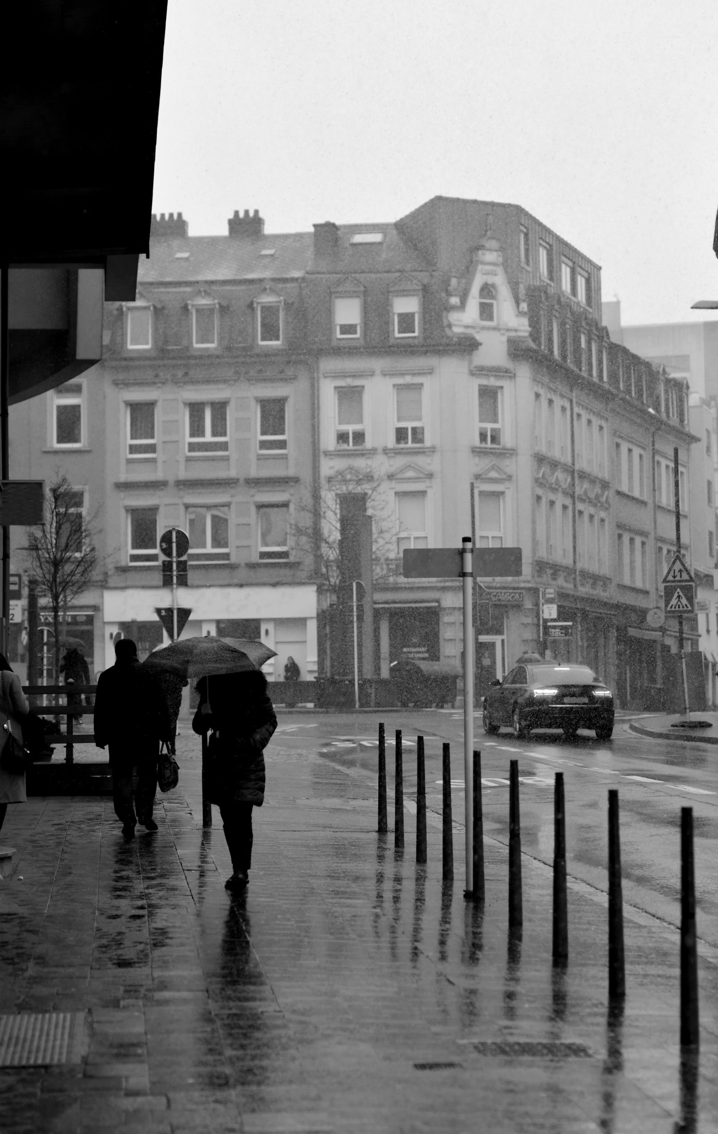 People walking with umbrellas on a rainy city street, with buildings and a parked car in the background.