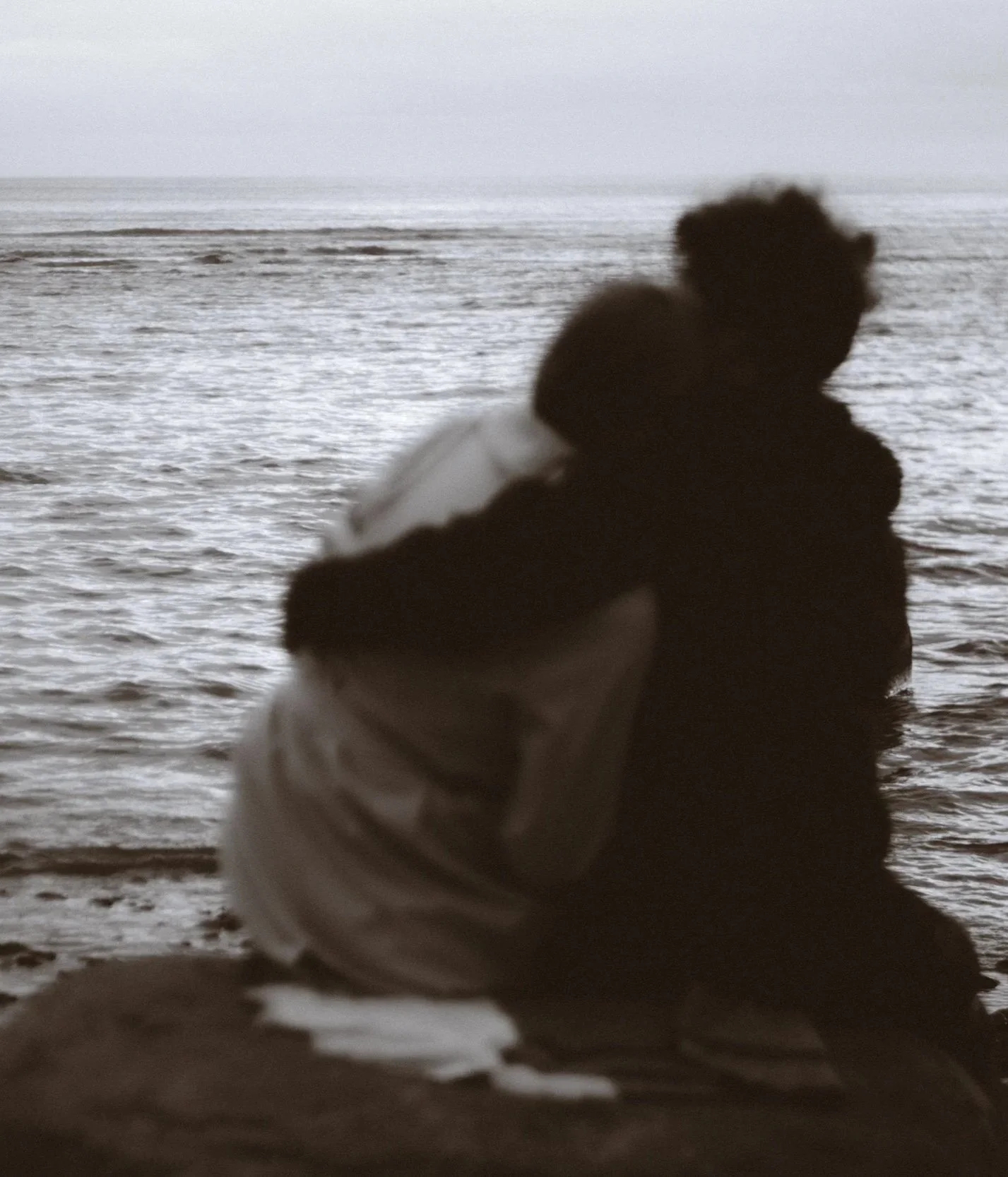 Silhouette of a person sitting on a rock by the ocean, with waves and a misty horizon in the background.