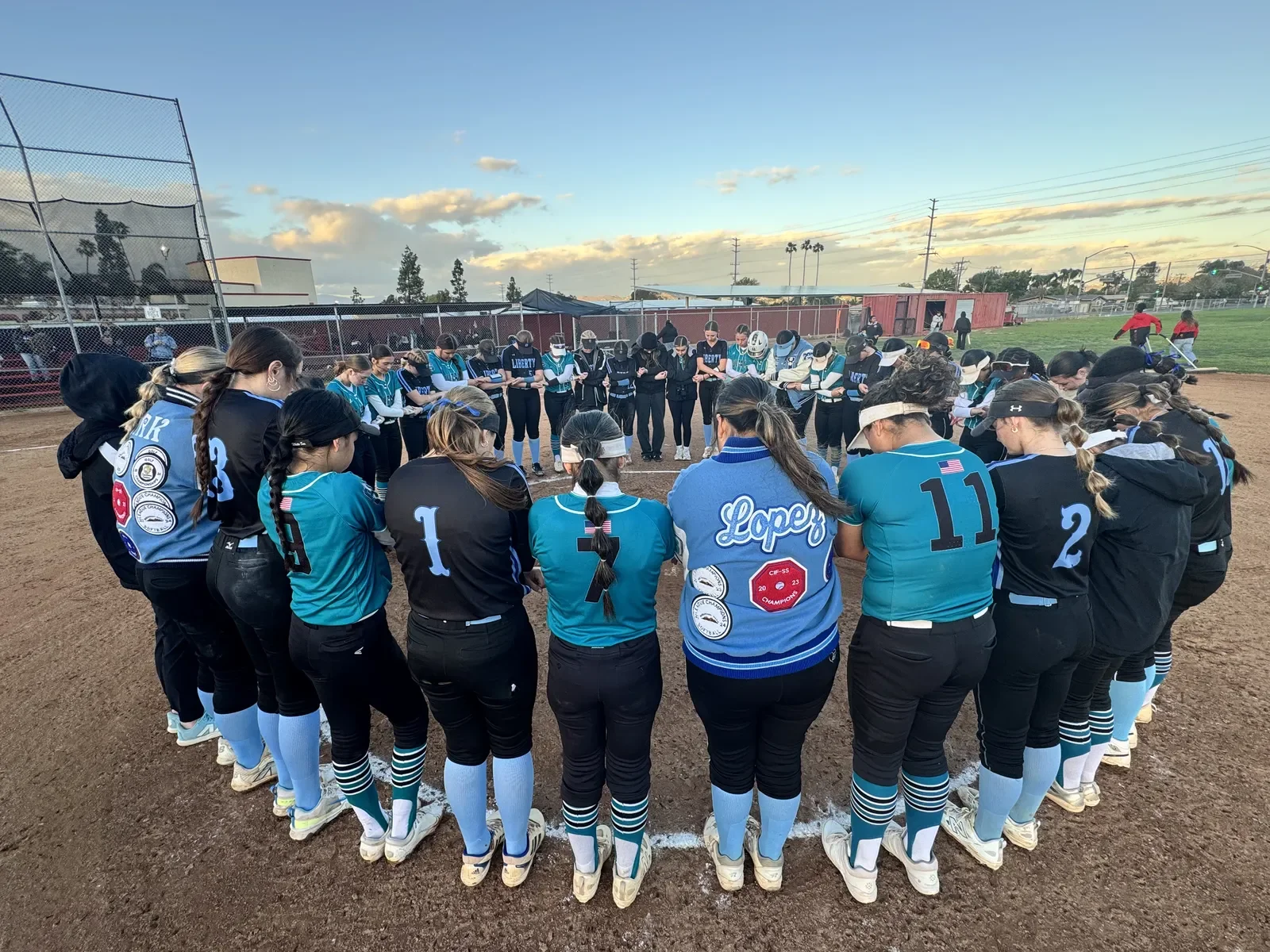 Softball team gathered in a circle on the field during sunset, praying before the game or practice.