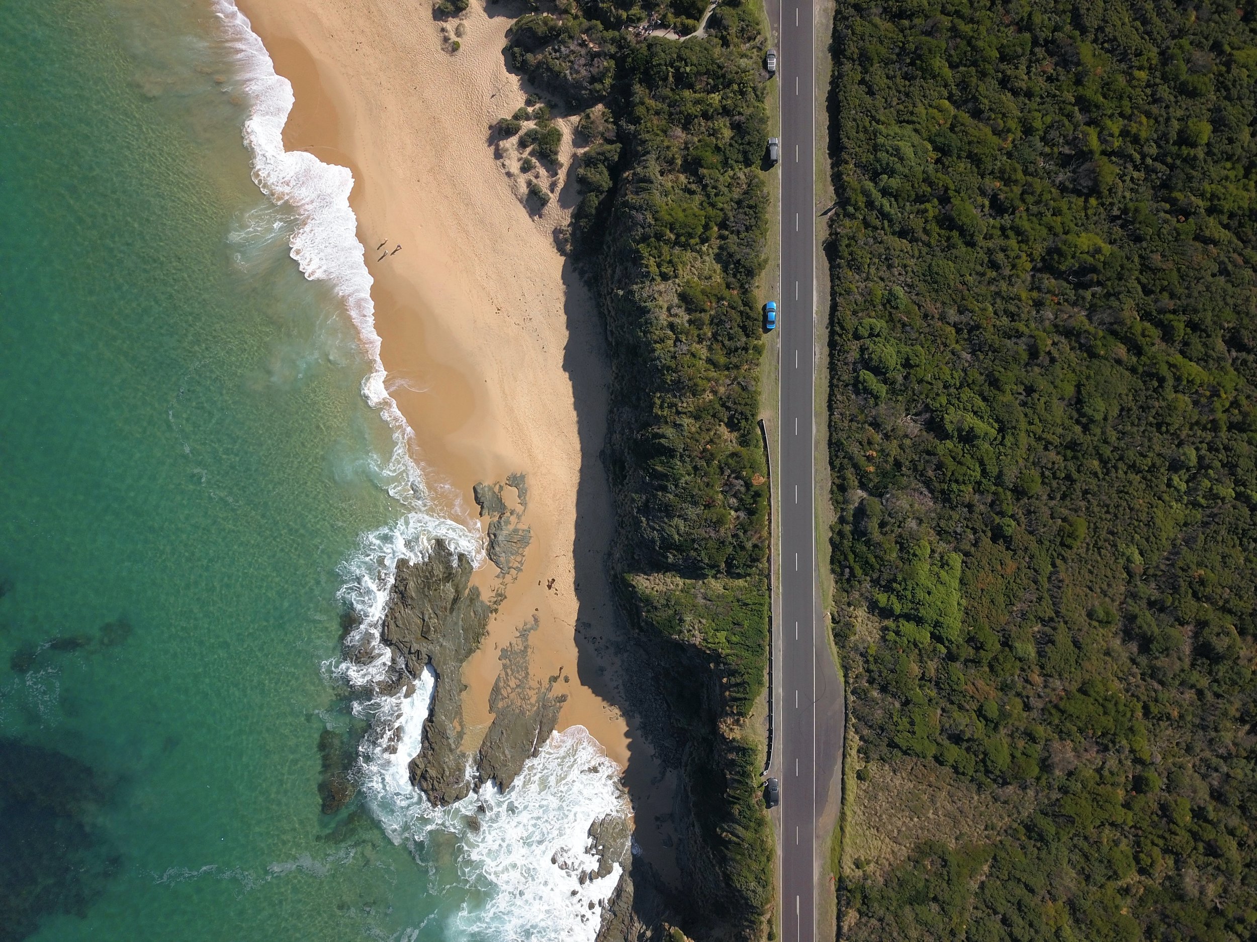 Aerial view of a beach with waves crashing on the sand, adjacent to a coastal road and dense greenery.
